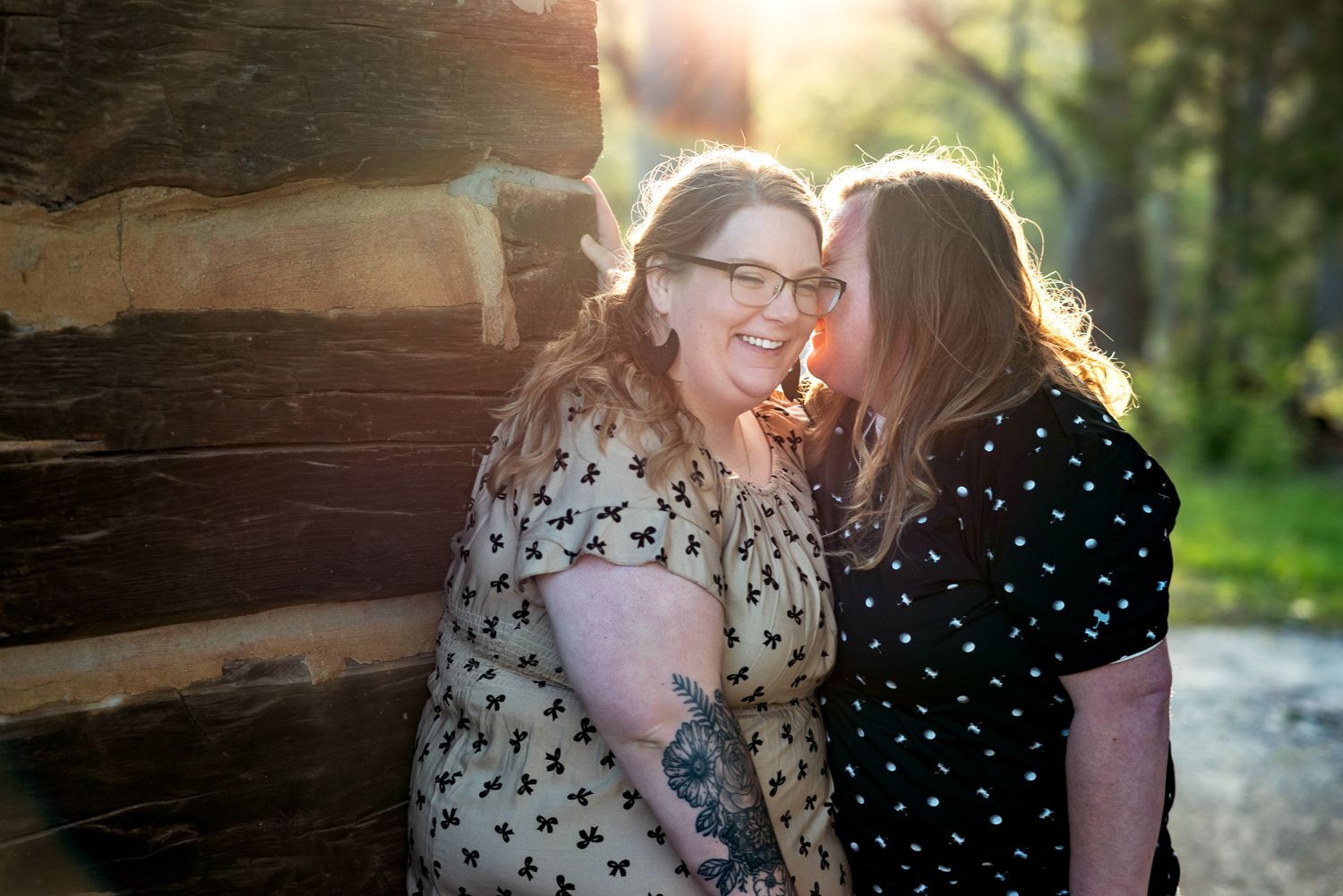 Two people share a joyful moment while standing against a rustic wooden wall in warm sunlight.
