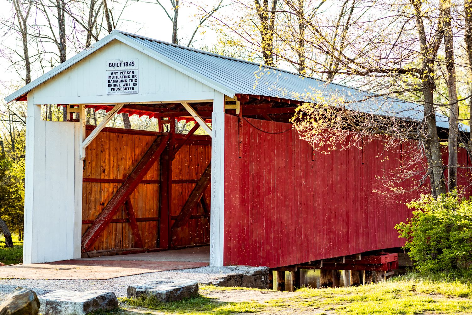 A white and red covered bridge with wooden doors stands among autumn trees in a rural setting.
