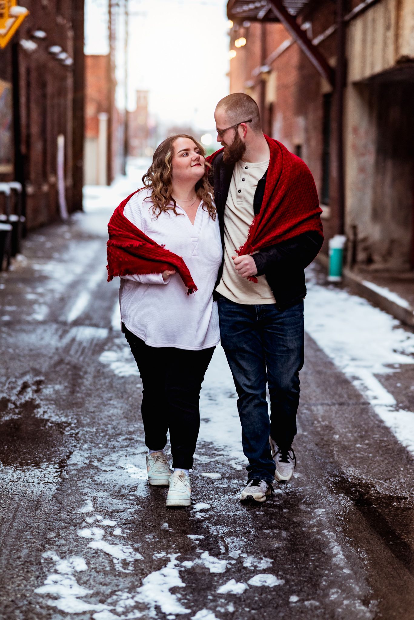 A couple in casual winter clothing sharing an intimate moment while walking down a snowy urban alley.