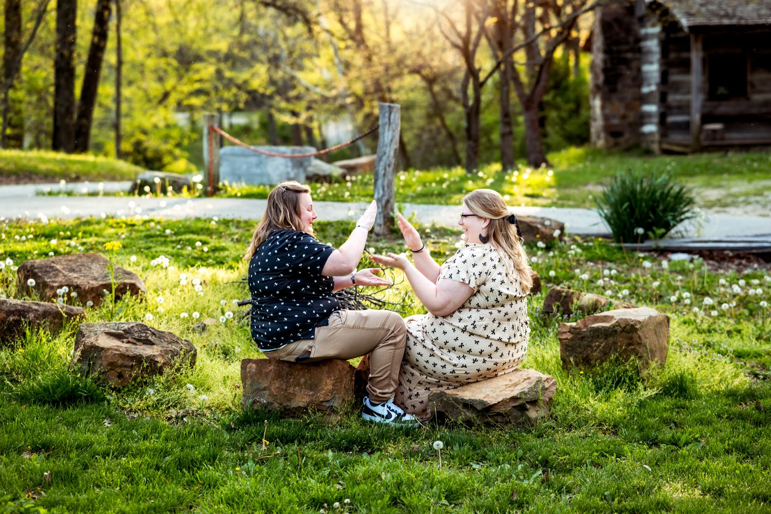 Two people sit on wooden stumps outdoors enjoying a warm afternoon at a rustic log cabin property.