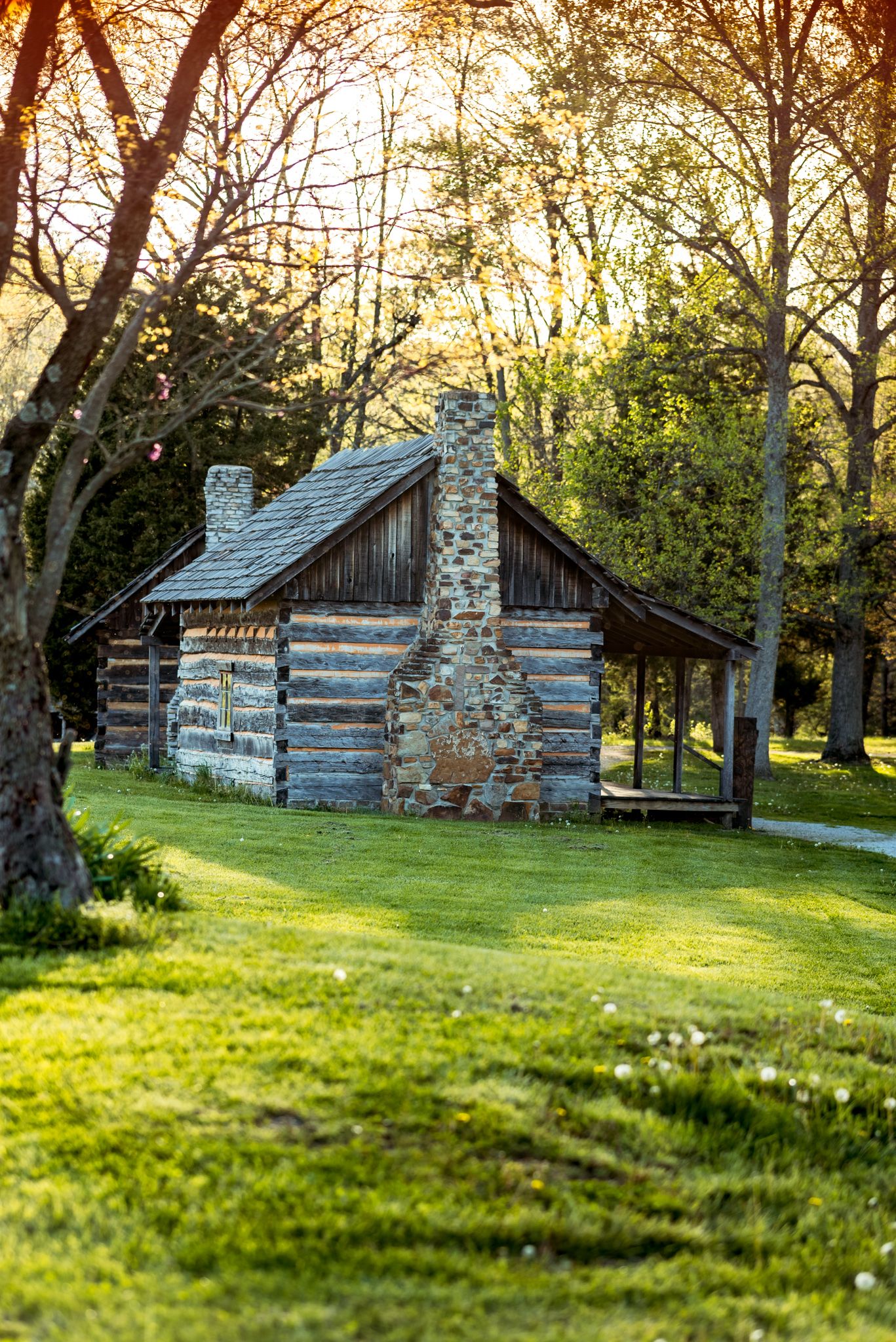 Rustic log cabin with stone chimney sits on lush green lawn at sunset, surrounded by tall trees in golden light.