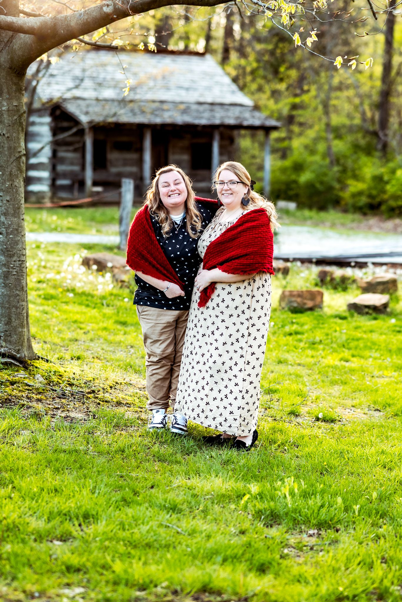 Two people in polka dot dresses and red shawls share a laugh by a rustic log cabin.