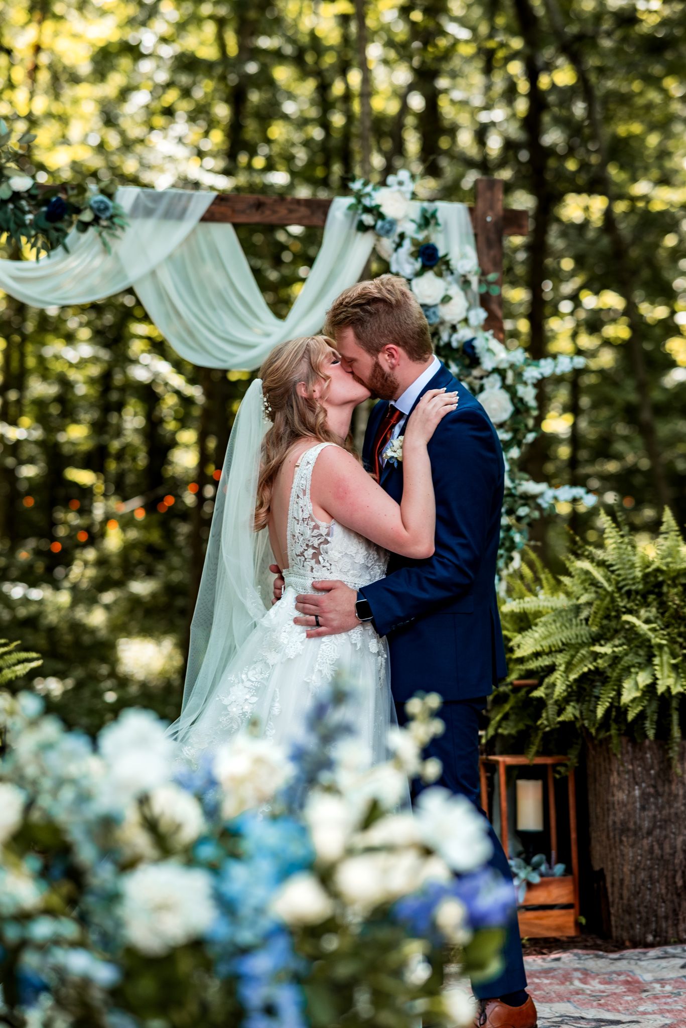 A romantic outdoor wedding ceremony moment under a white draped arch with blue flowers and greenery in a wooded setting.