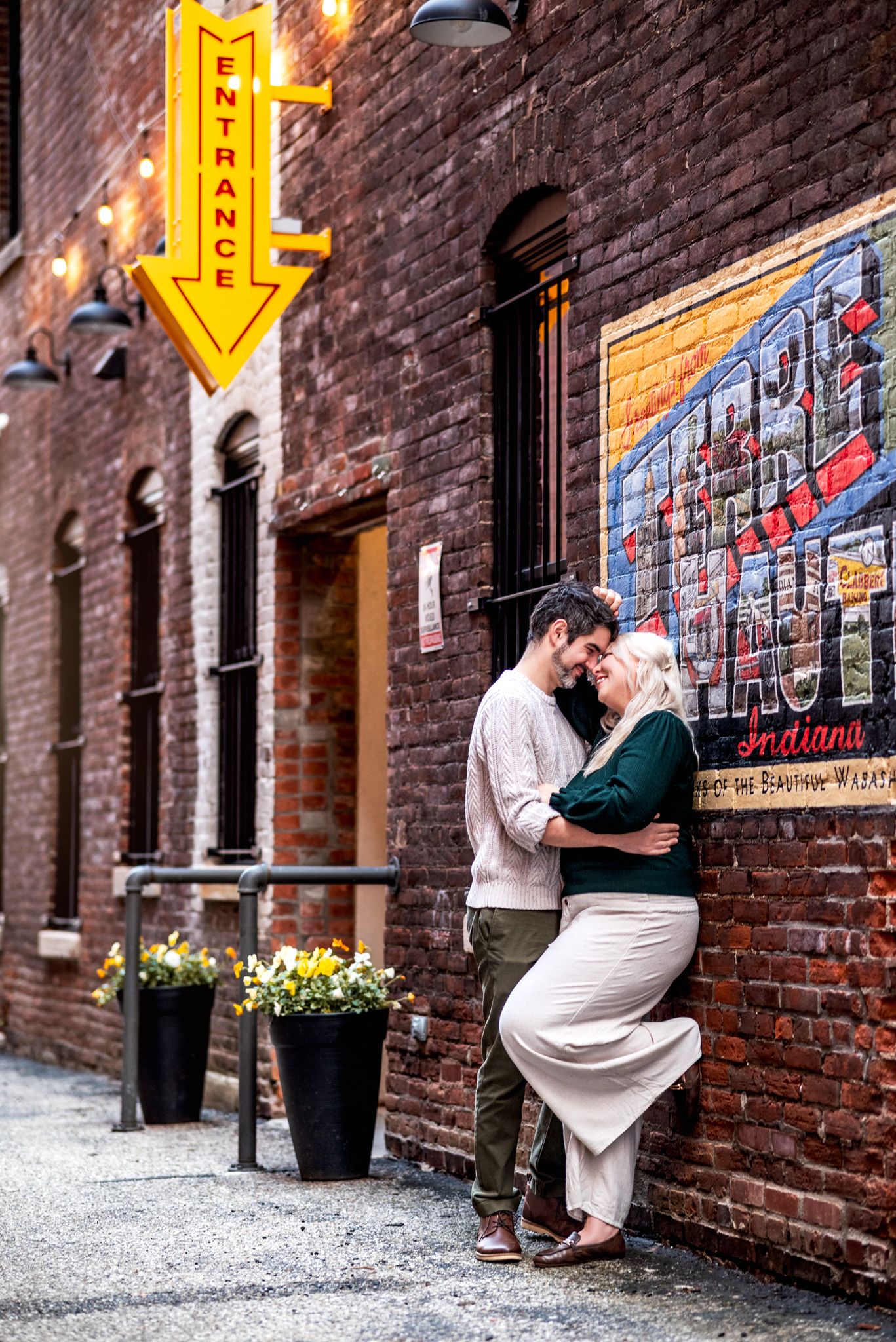 Couple embraces against brick wall in urban alley with yellow arrow sign and colorful mural artwork visible.