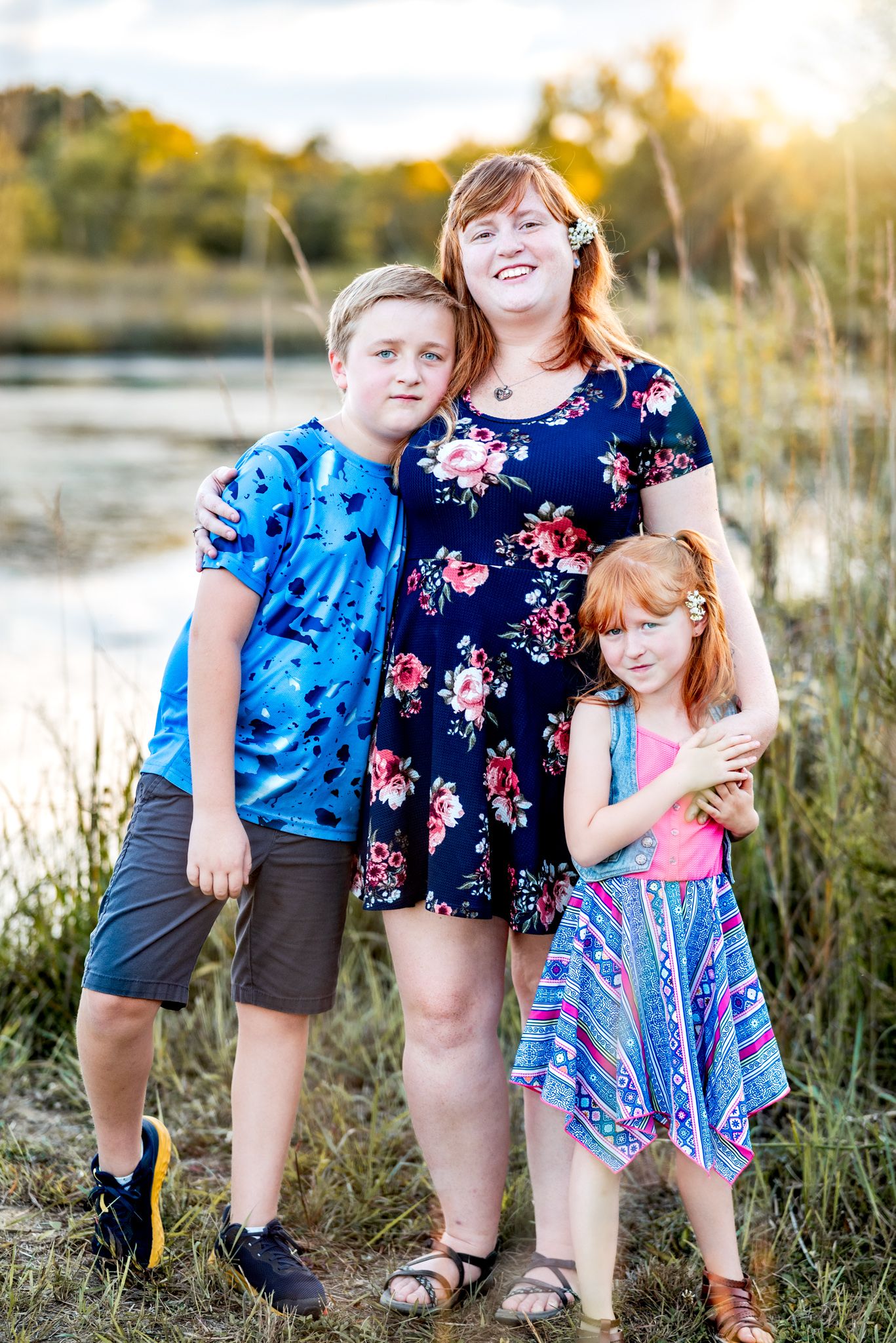 A family poses together outdoors at sunset near a lake wearing colorful patterned clothing on a summer evening.