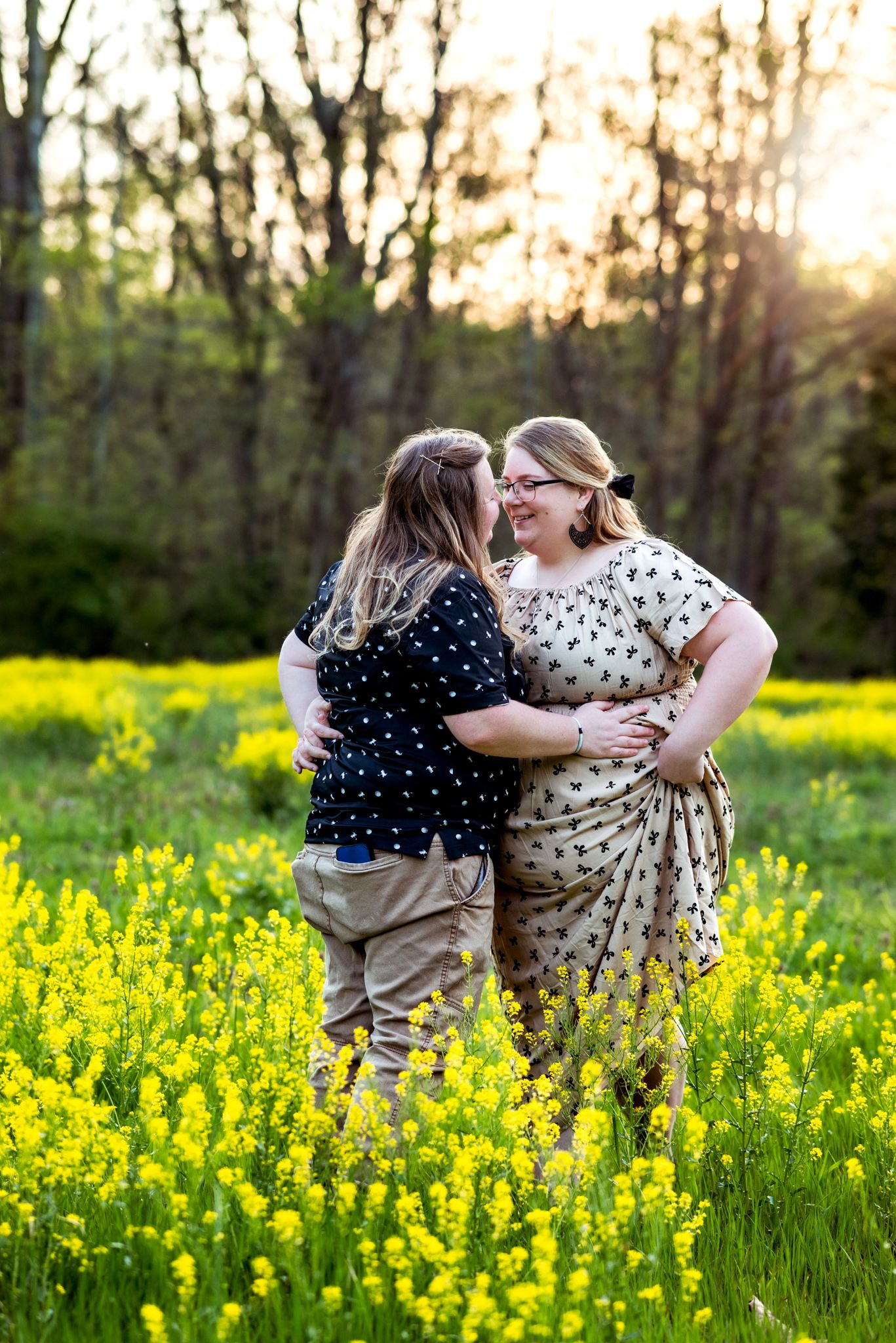 Two figures embrace while standing in a vibrant field of yellow wildflowers at sunset with trees in the background.