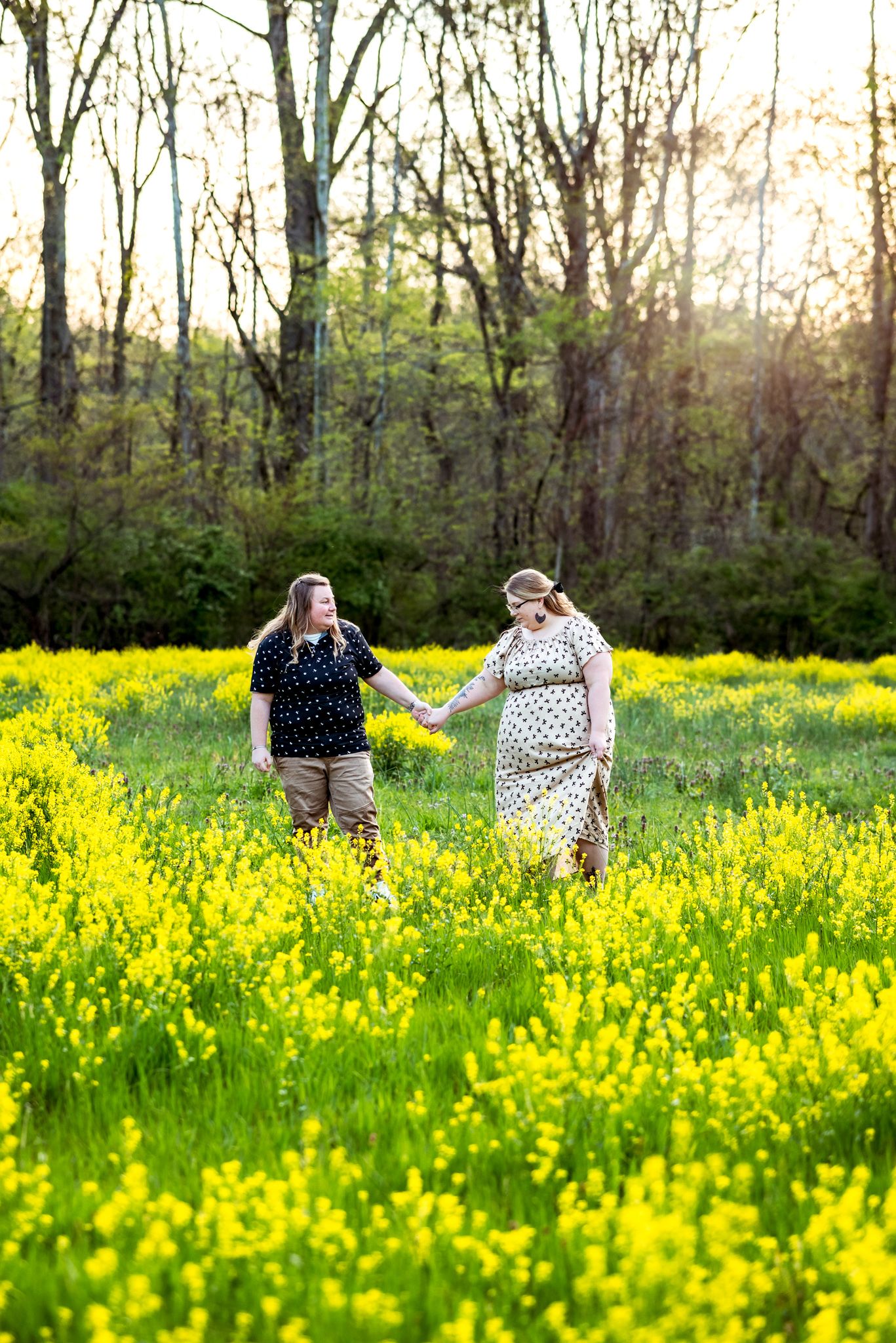 Two people walk hand in hand through a vibrant field of yellow wildflowers at sunset with trees in the background.