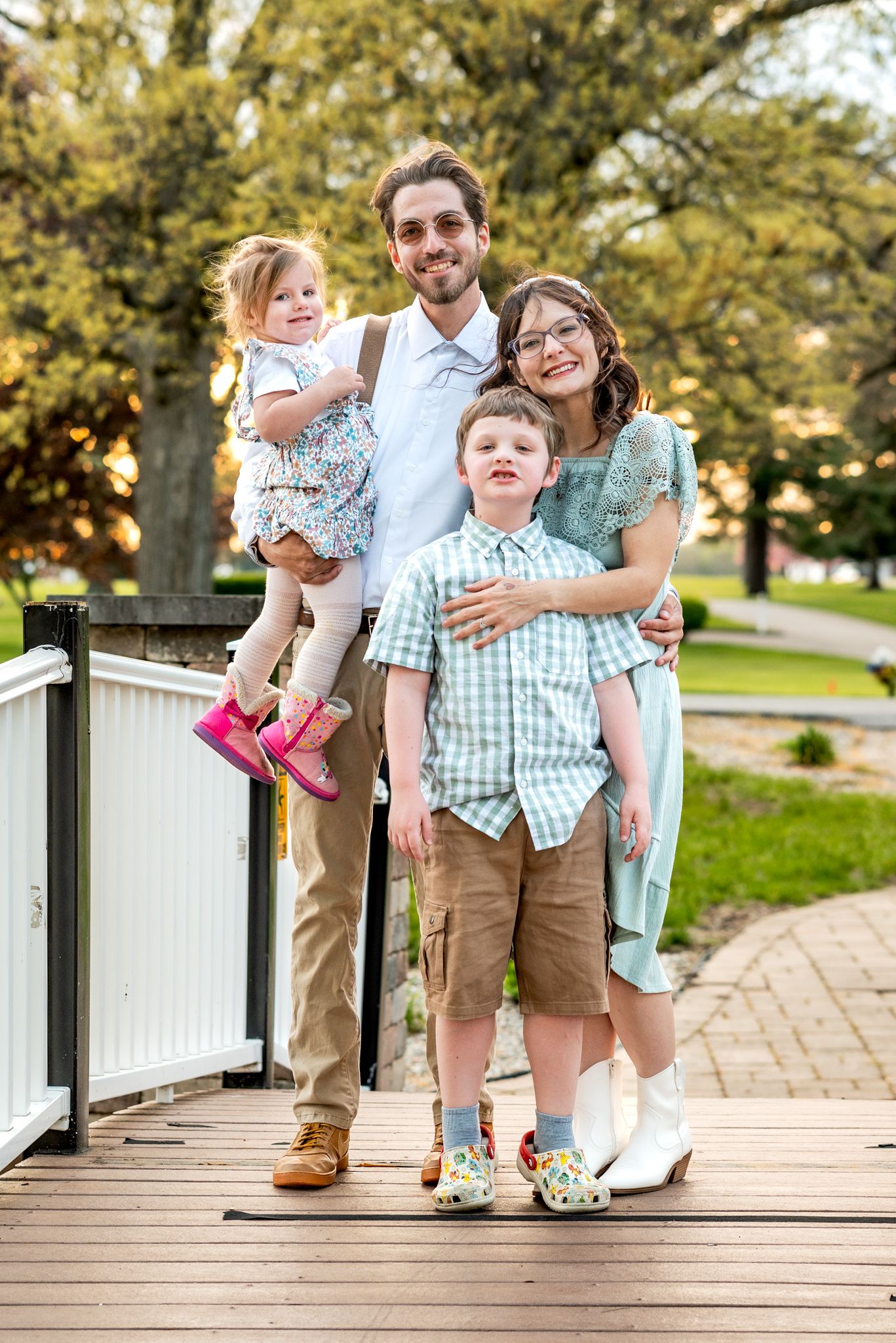 A family of four poses together on a wooden deck surrounded by green trees on a sunny day.