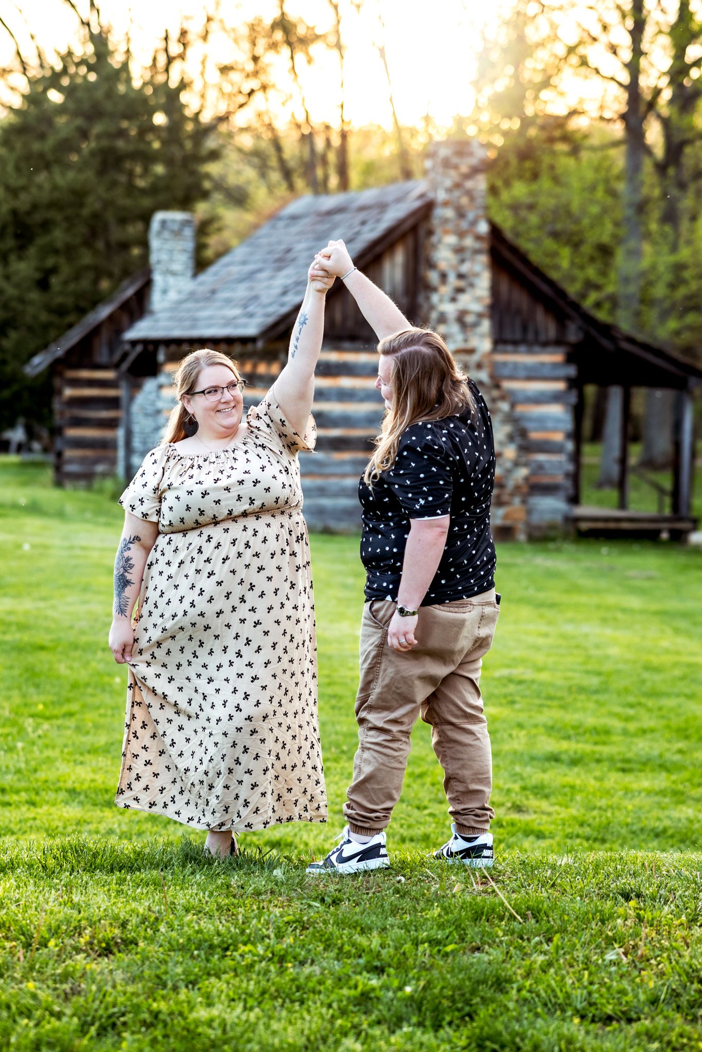A couple dances together on a green lawn with a rustic log cabin in the background at sunset.
