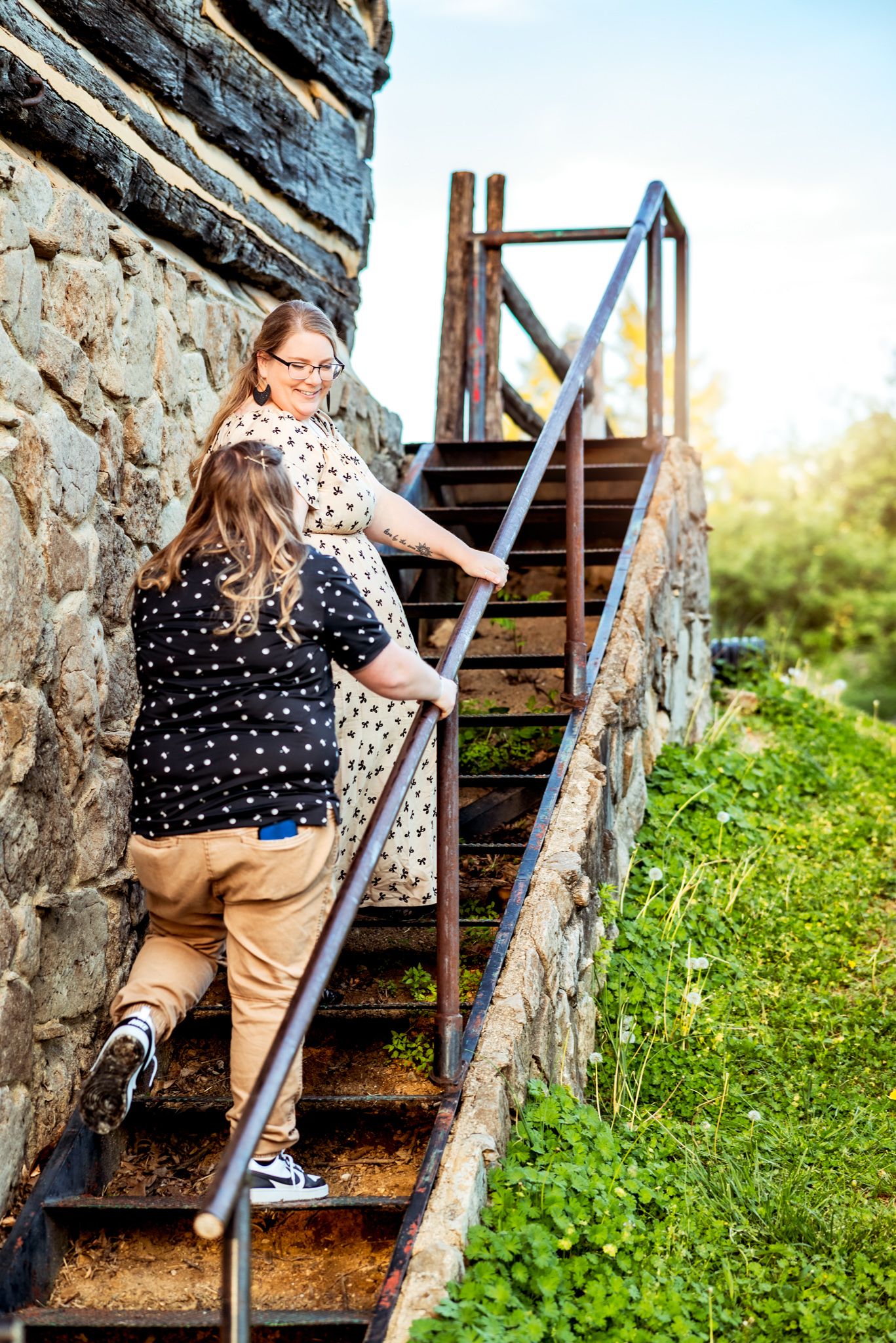 Two people climb rustic metal stairs against a stone wall surrounded by green grass and natural scenery.