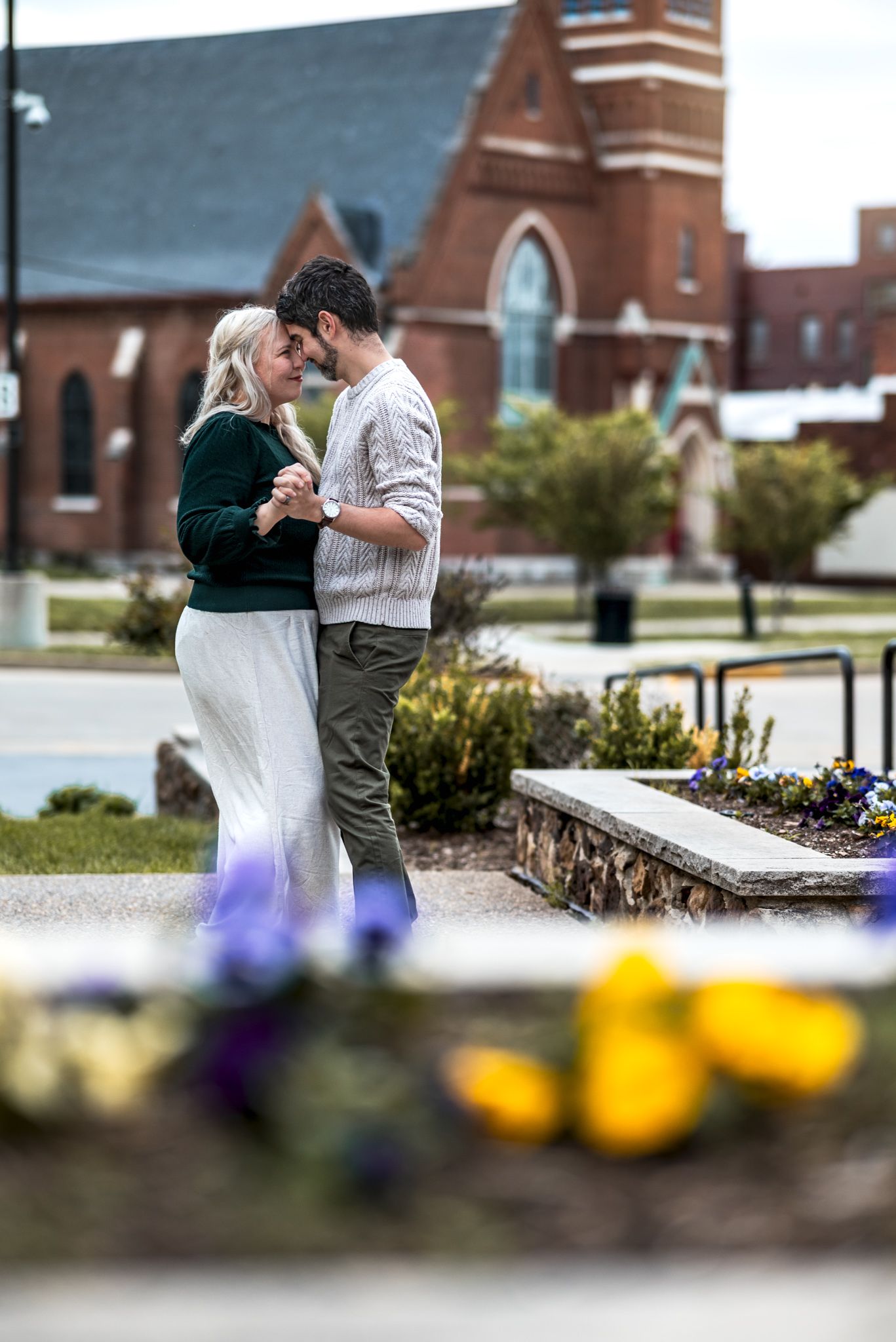 Romantic couple shares affectionate moment outdoors while sitting on stone wall with colorful flowers in foreground.