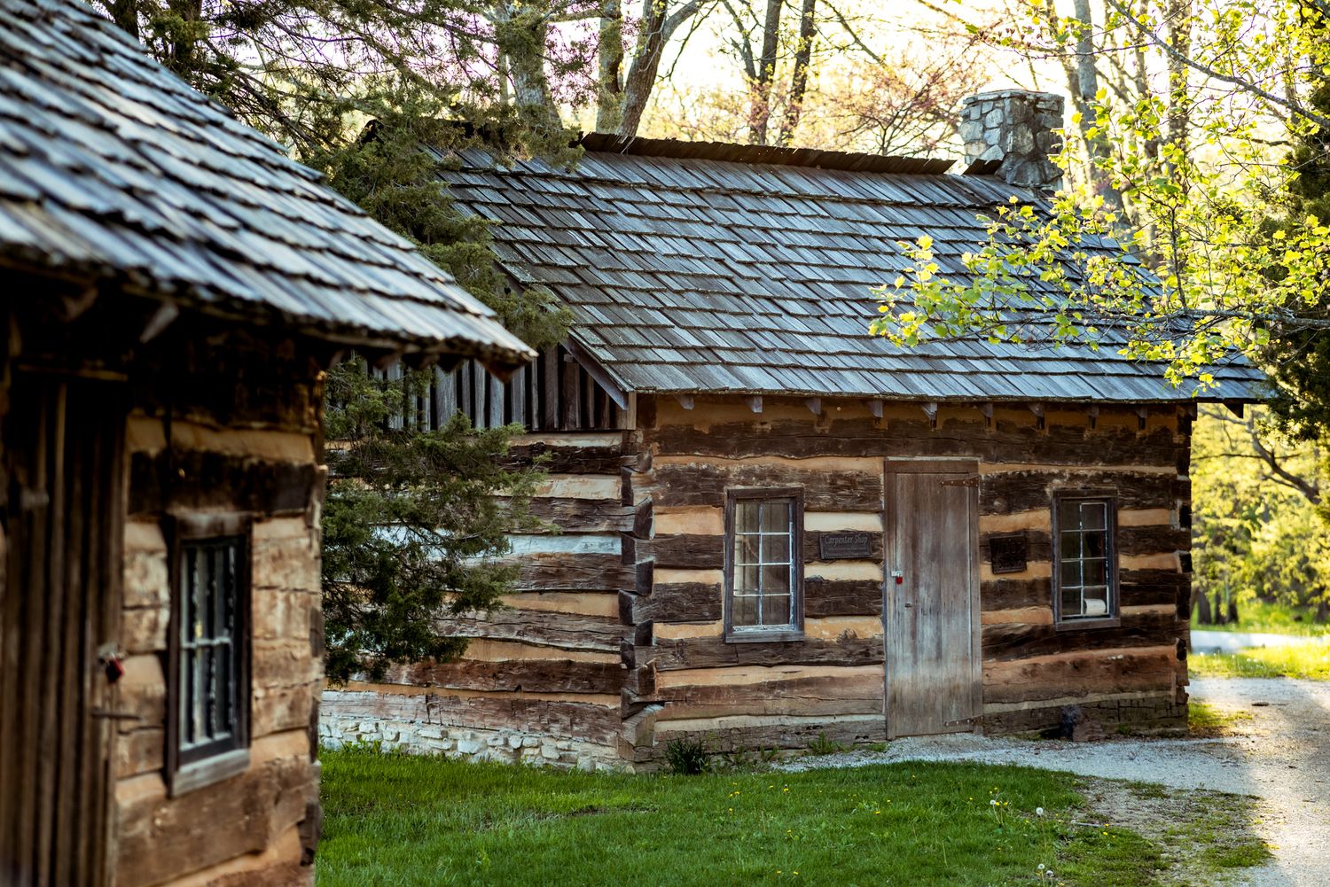 Historic rustic log cabin with weathered wooden walls and slate roof nestled among trees in a tranquil woodland setting.