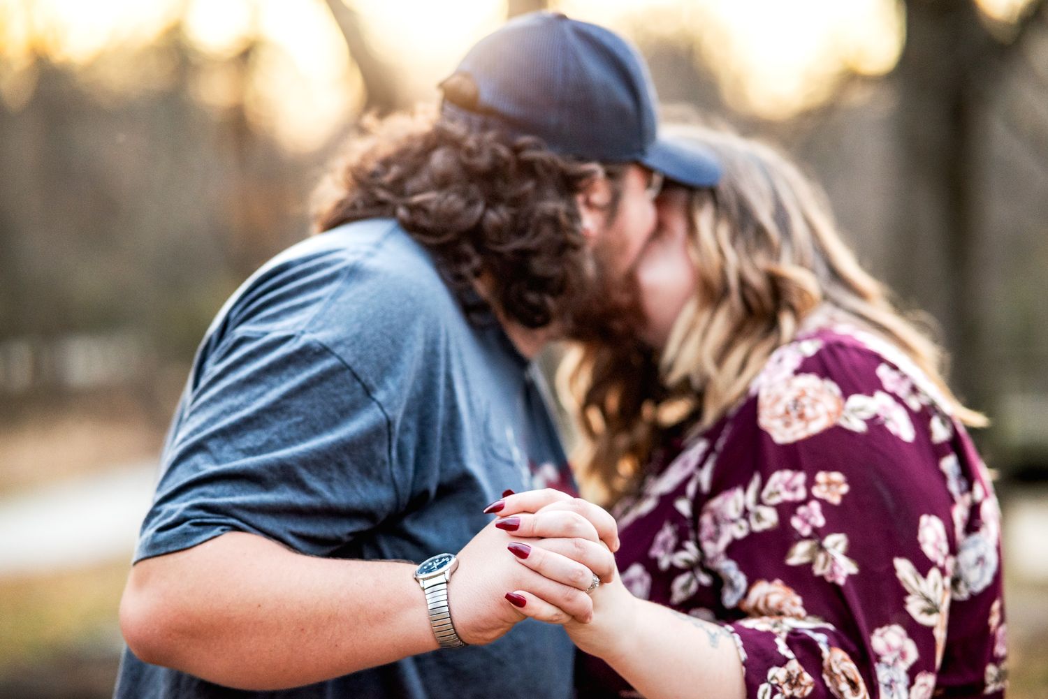 A romantic couple in burgundy and blue outfits embrace during a golden sunset photoshoot in a park setting.