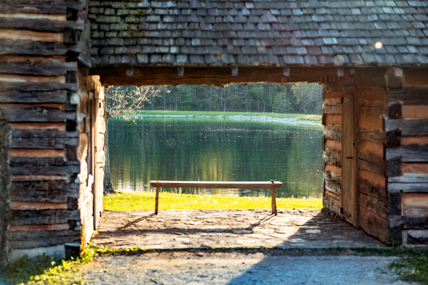 View through a rustic log cabin doorway framing a serene lake with sunlit grass and wooden fence in the foreground.