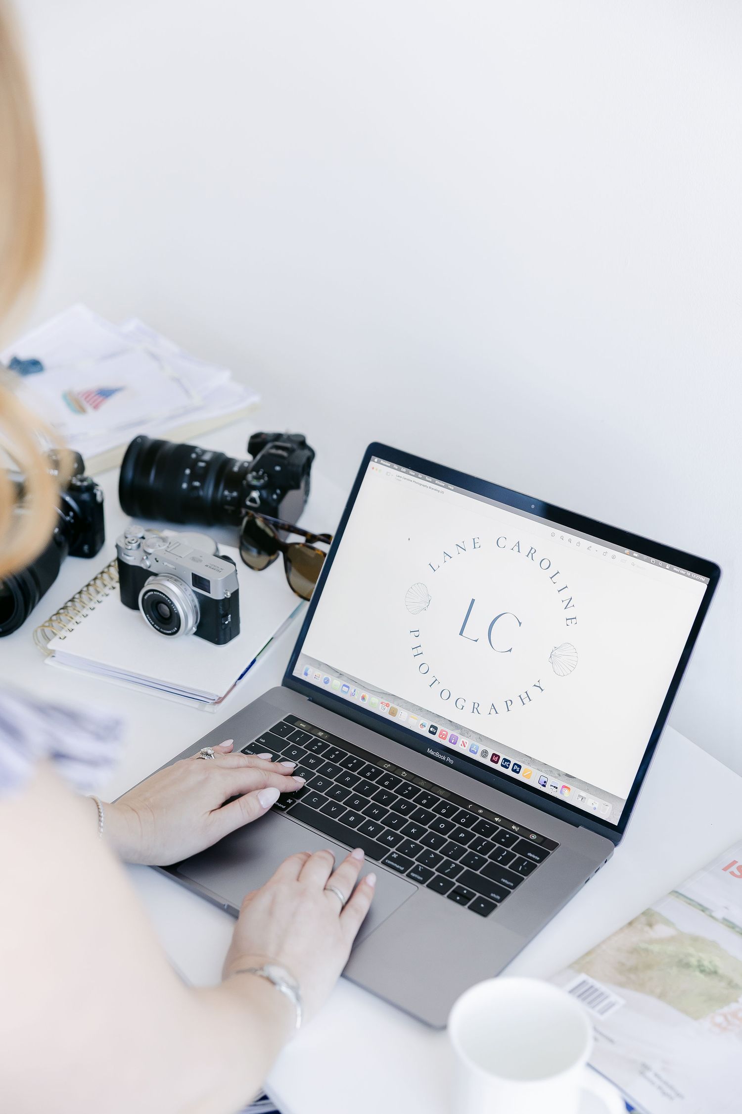 Person working at white desk with MacBook laptop, camera equipment and coffee cup in minimalist workspace setup.