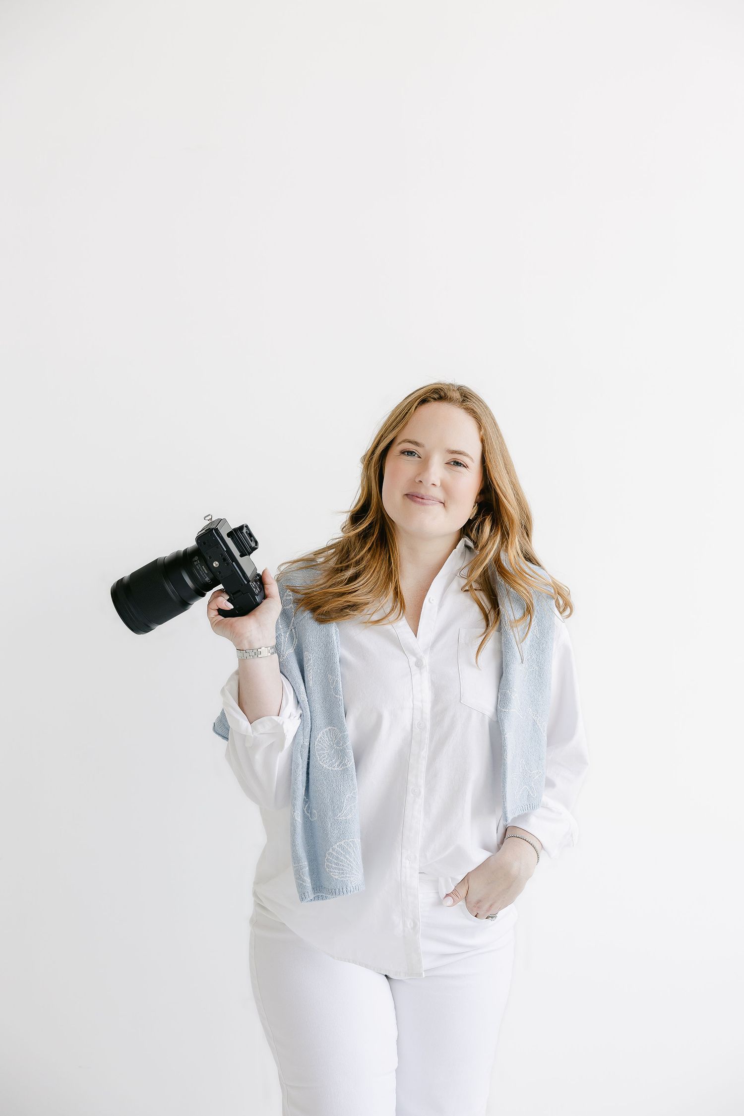 Professional photographer in white outfit holding camera against white background.
