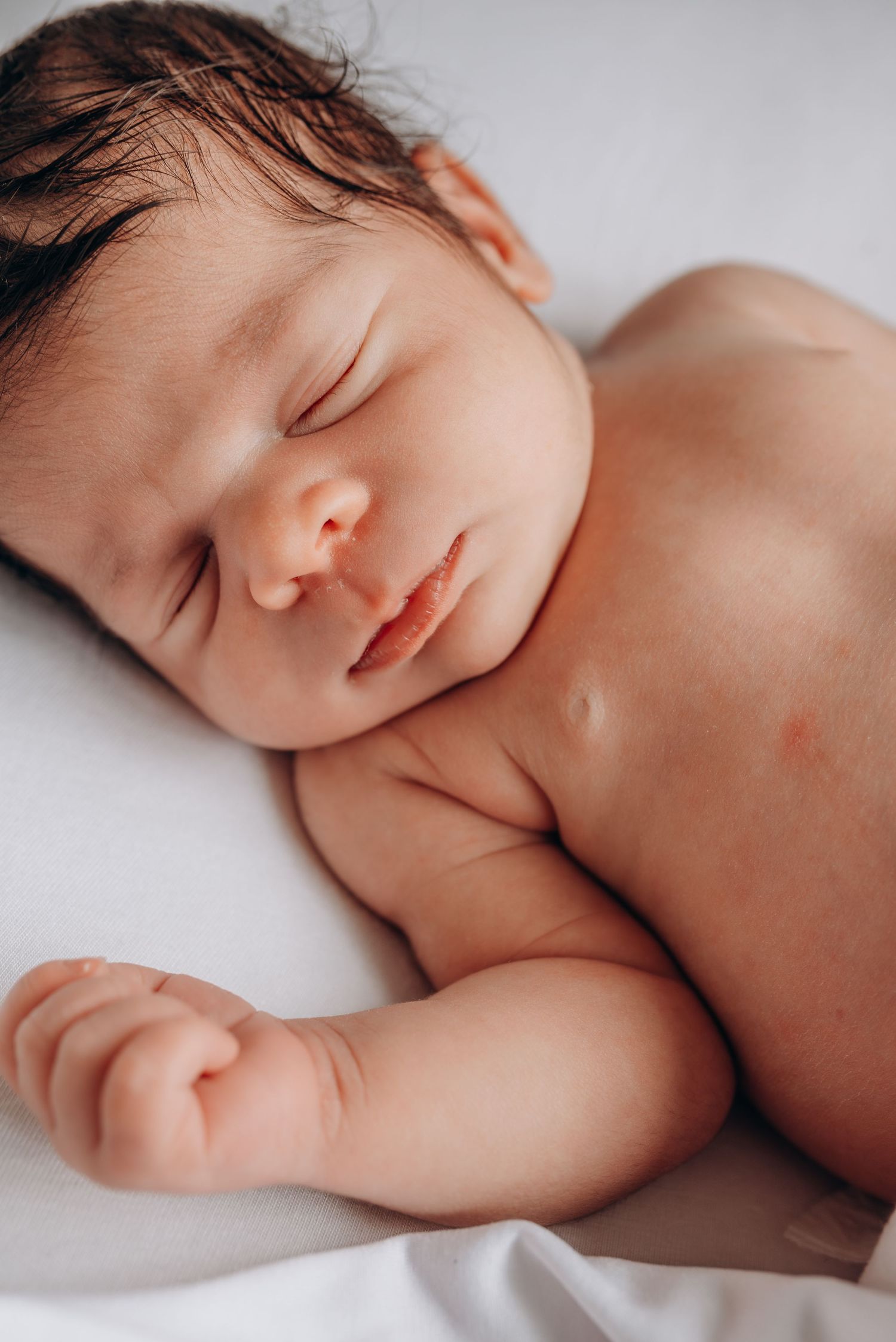A peaceful sleeping newborn baby with dark hair resting on white bedding in soft lighting.