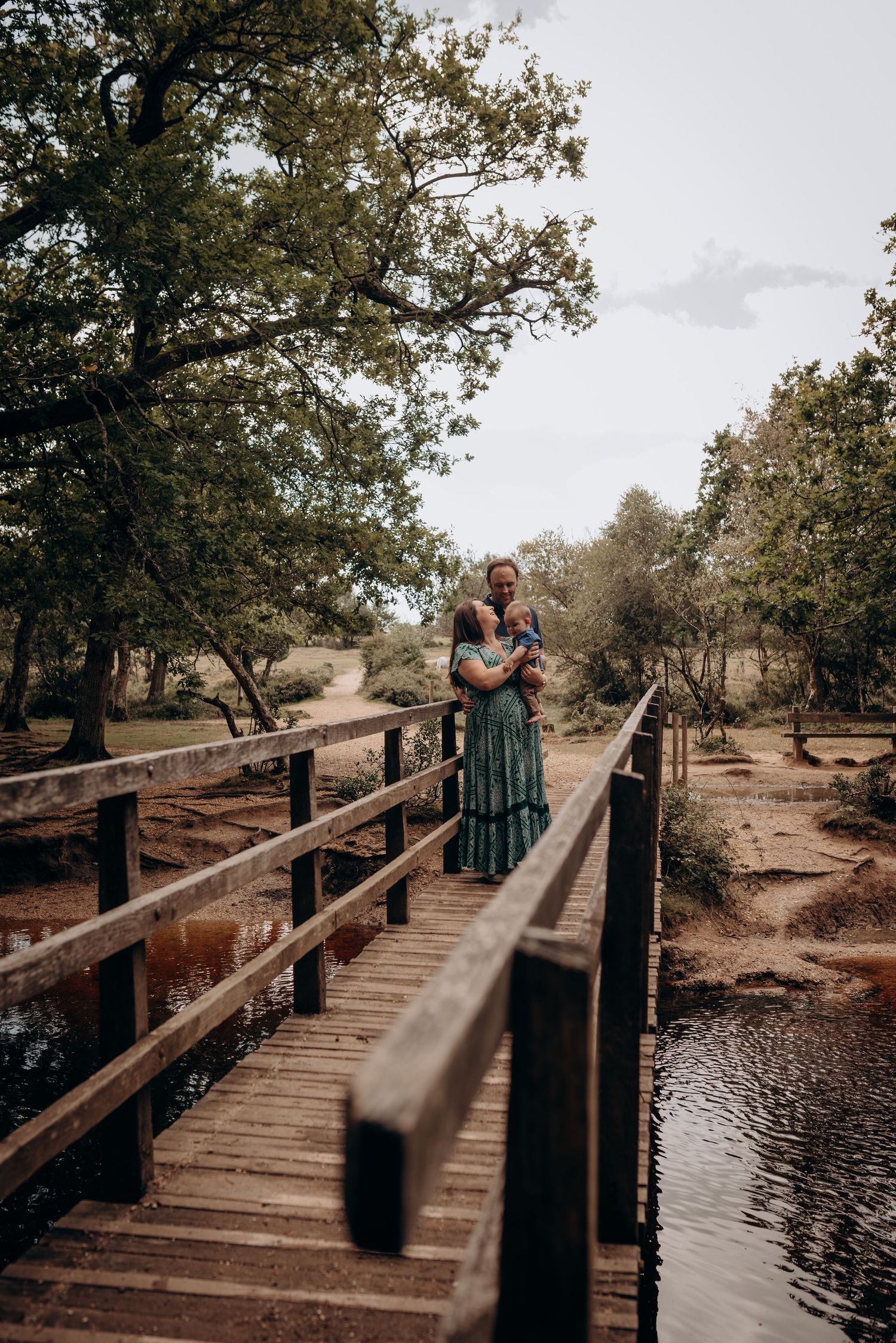 A couple shares an intimate moment on a wooden bridge over water surrounded by trees.