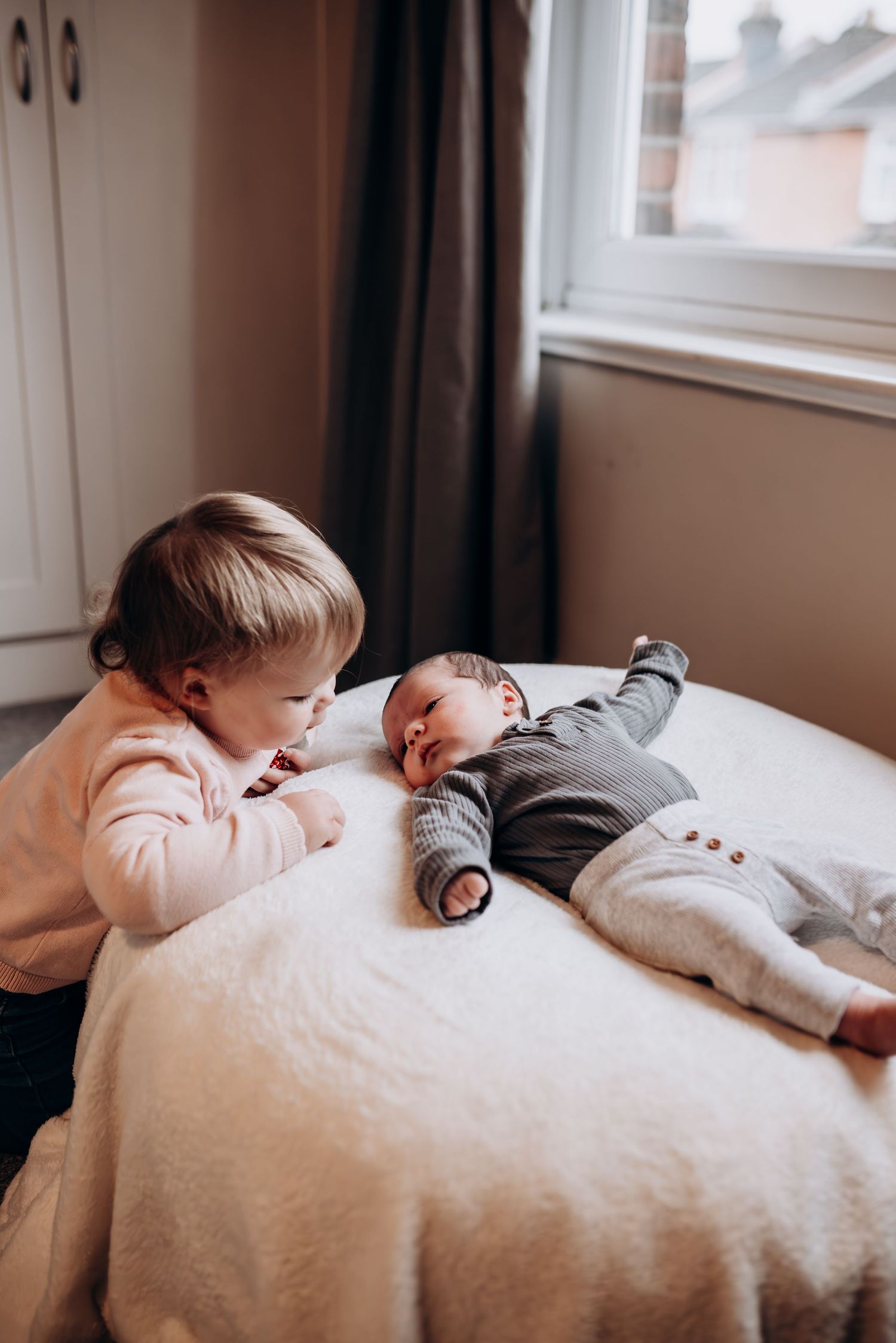 Siblings lying on bed together, showing gentle interaction with newborn baby.