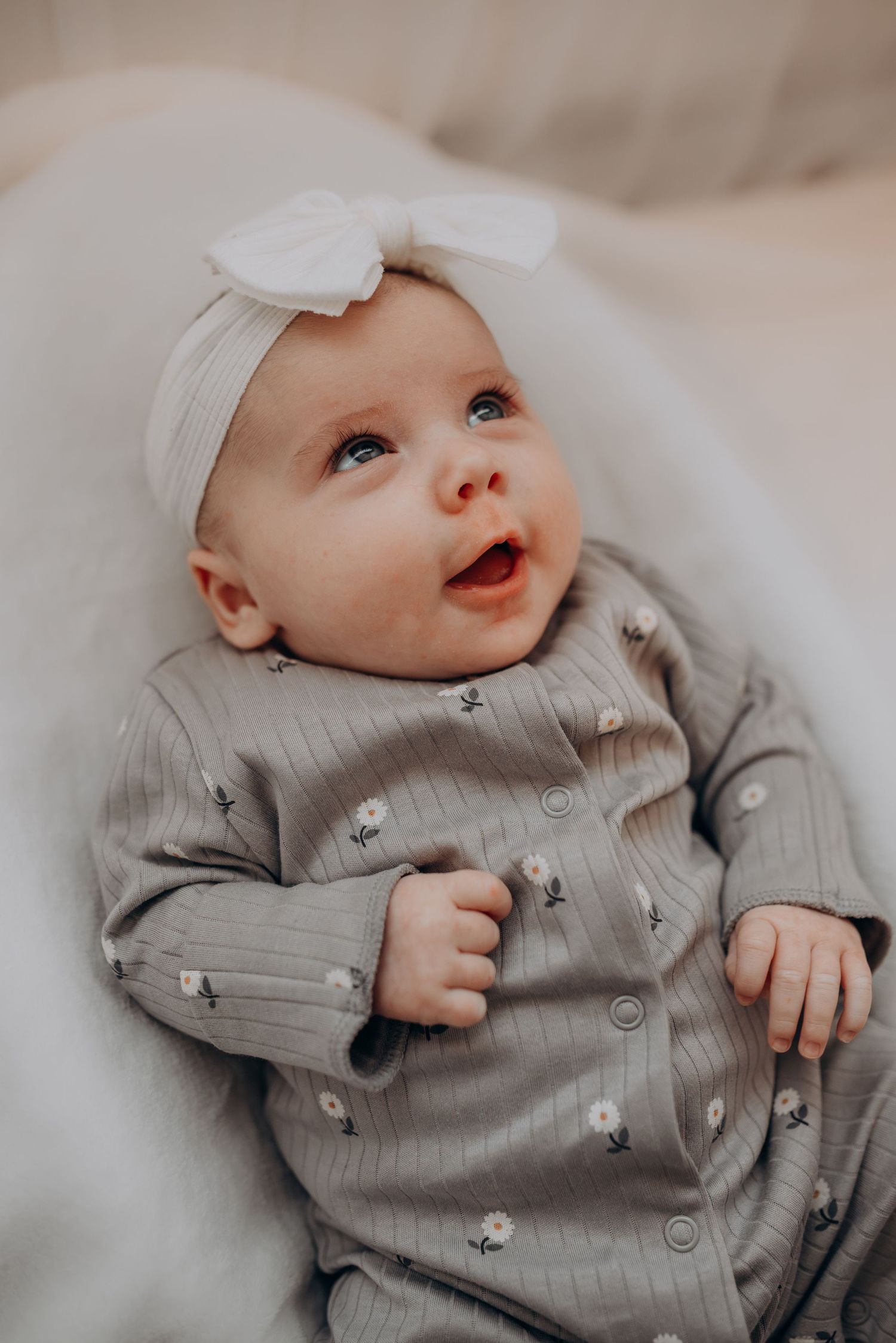 A baby in gray clothing with a white bow headband gazes upward with bright eyes.