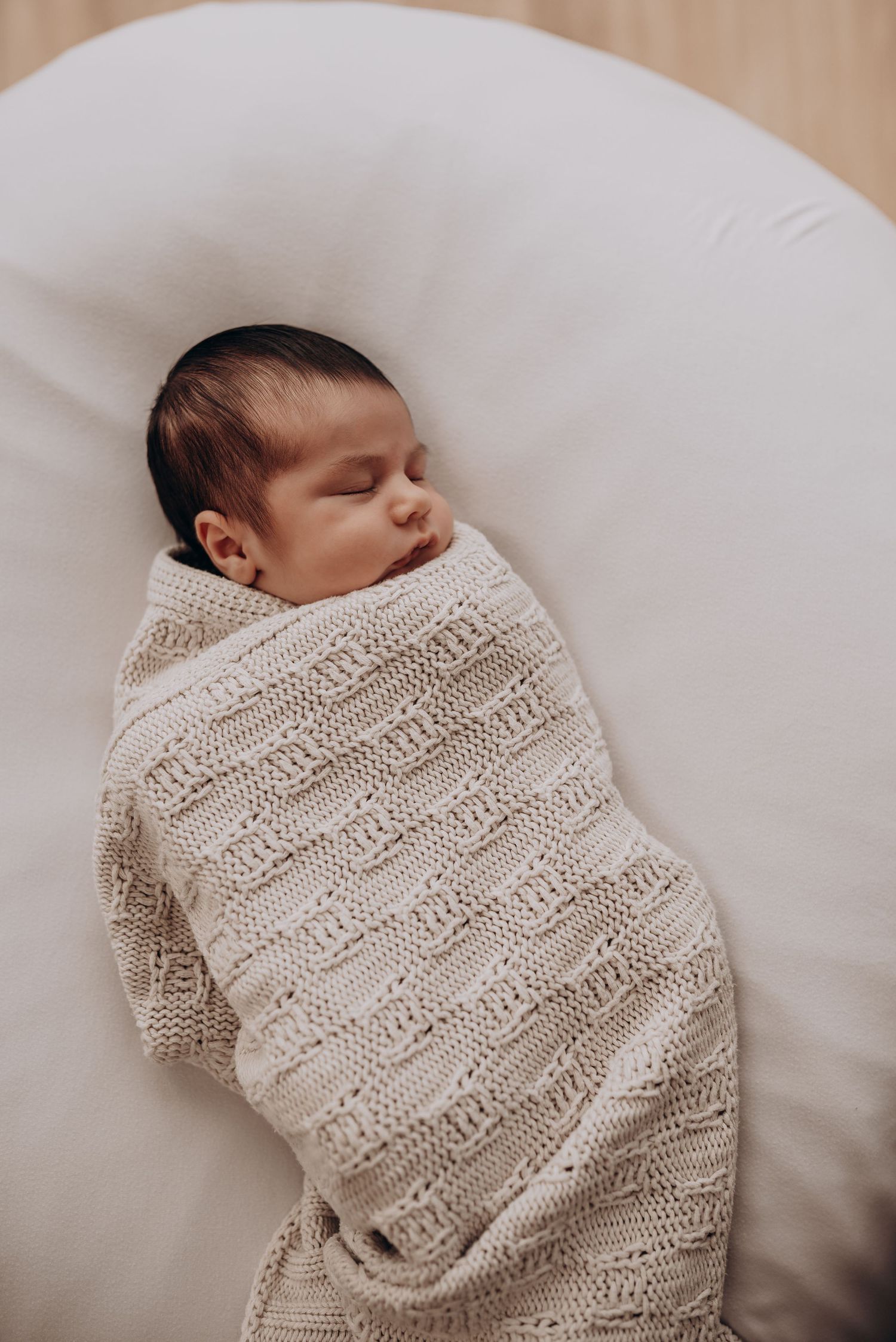 Newborn baby sleeping peacefully wrapped in cream knitted blanket on white bedding.