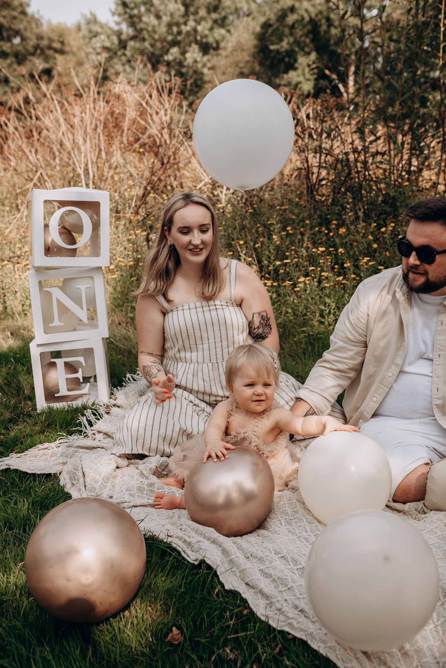Family picnic scene with white and gold balloons on blanket in grassy field.