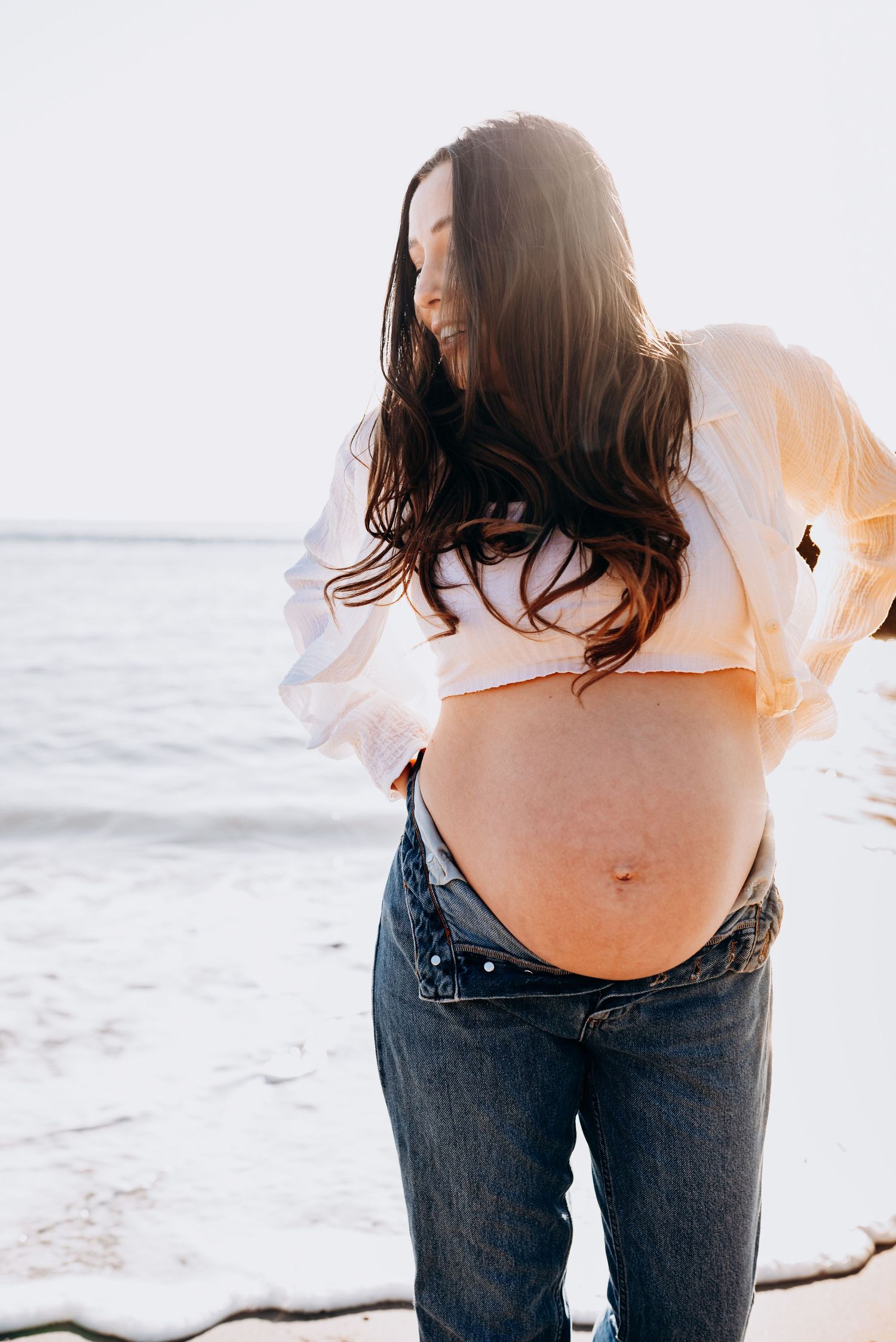 Maternity photo at beach showing pregnant belly with white top and jeans at sunset.