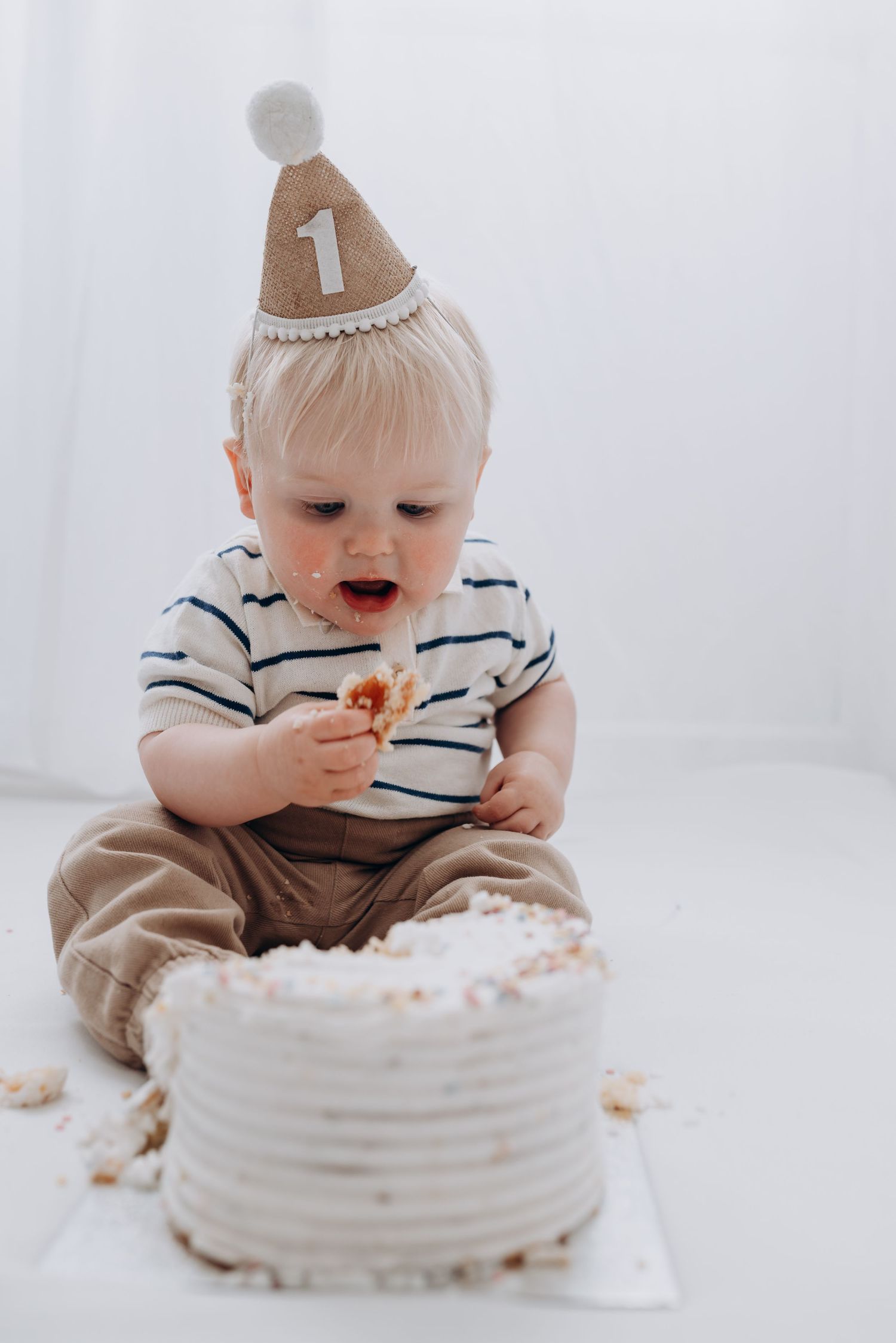 Baby in striped shirt enjoying their first birthday cake smash photoshoot with white frosted cake.