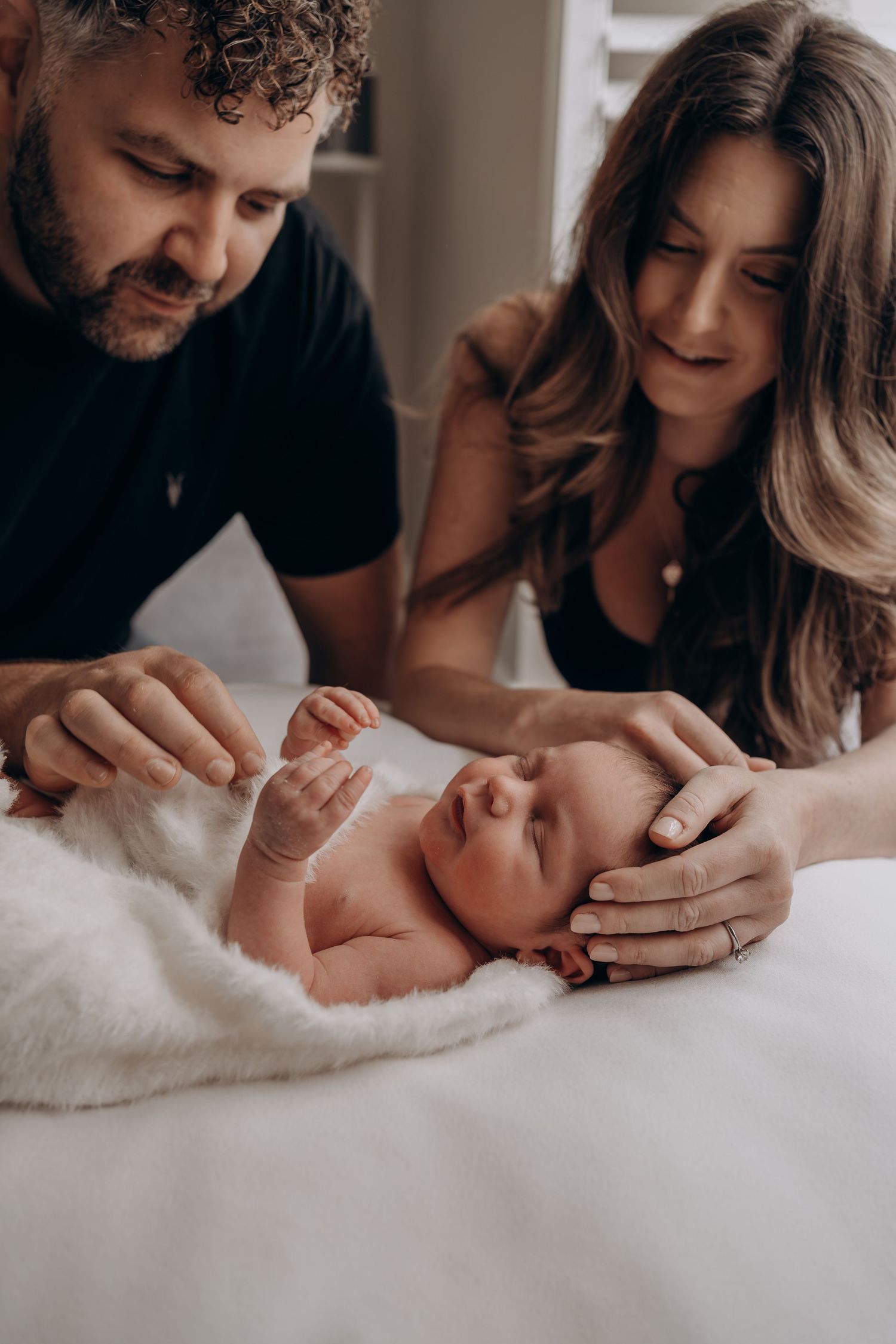 Parents tenderly gazing at their newborn baby lying on a white fluffy blanket on a bed.