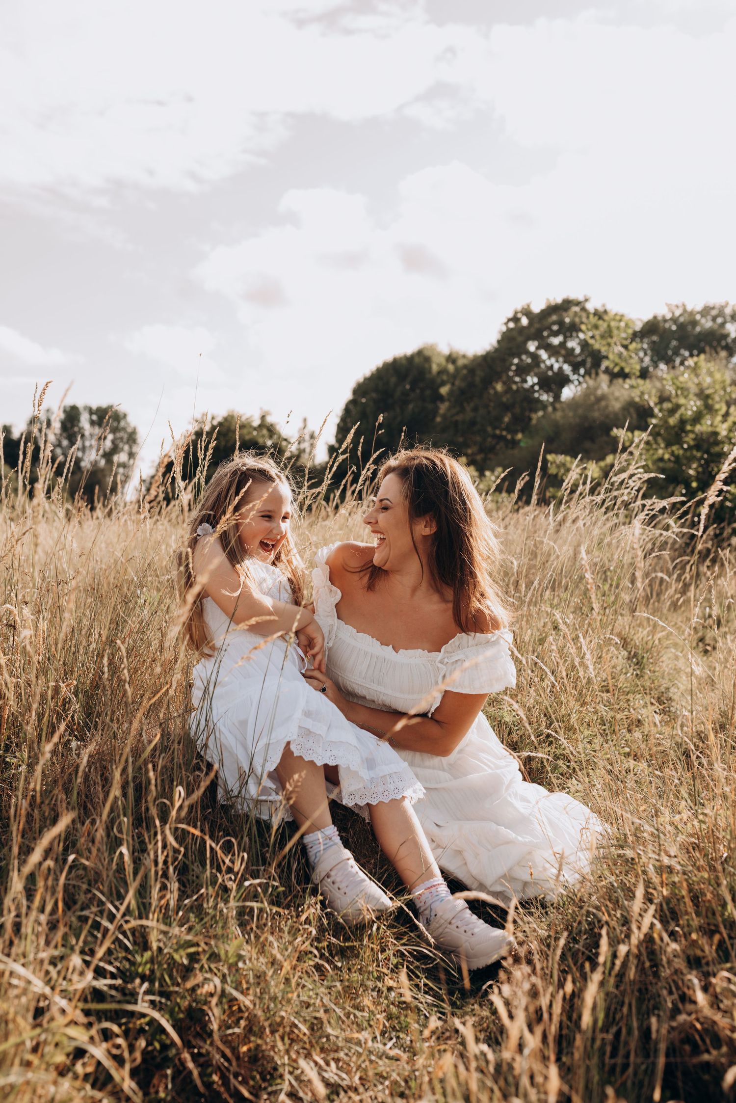 Mother and daughter in white dresses share joyful moments together in a golden sunlit field of tall grass.