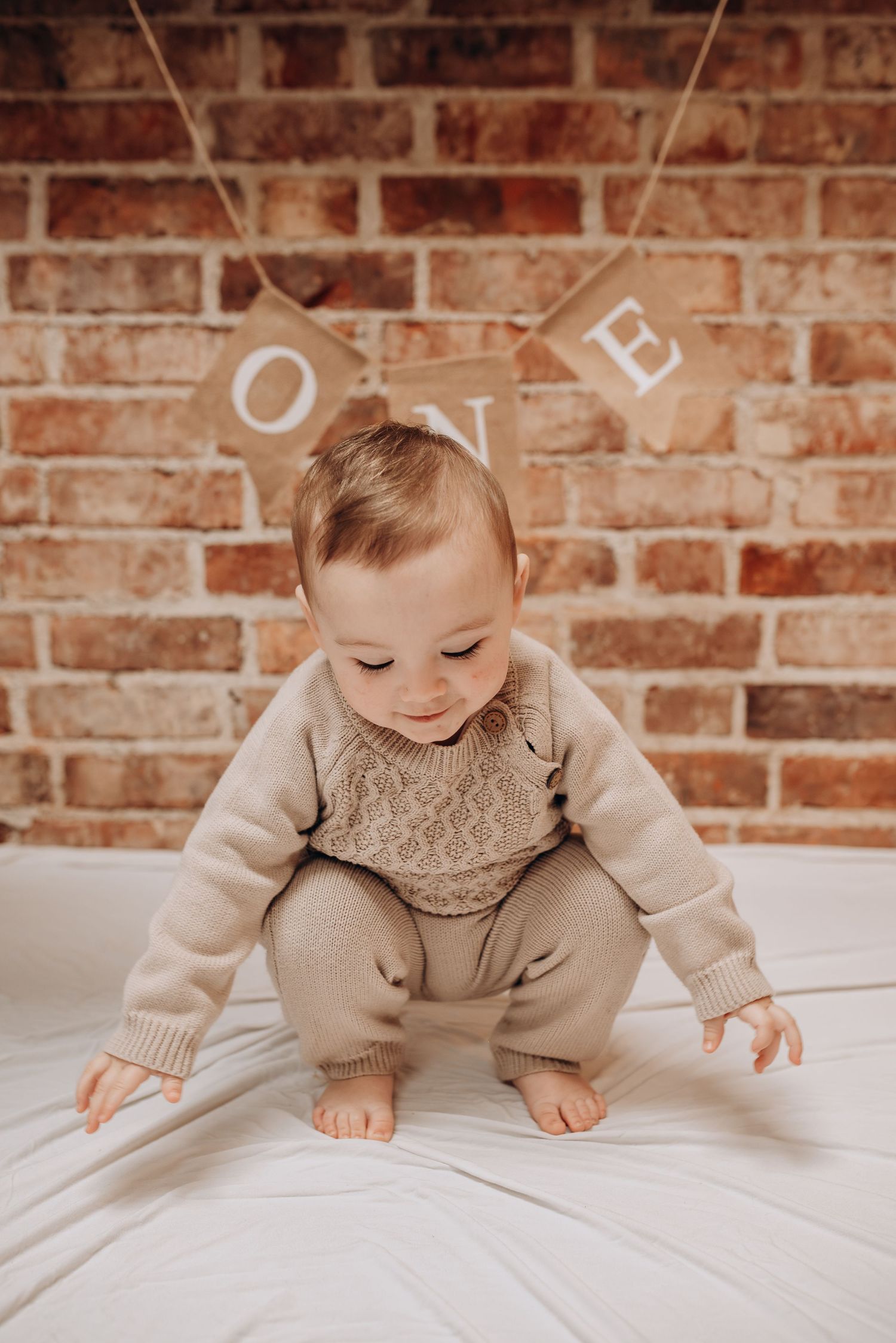 A baby in beige knitwear sits on white fabric with 'ONE' banner decoration against brick wall.