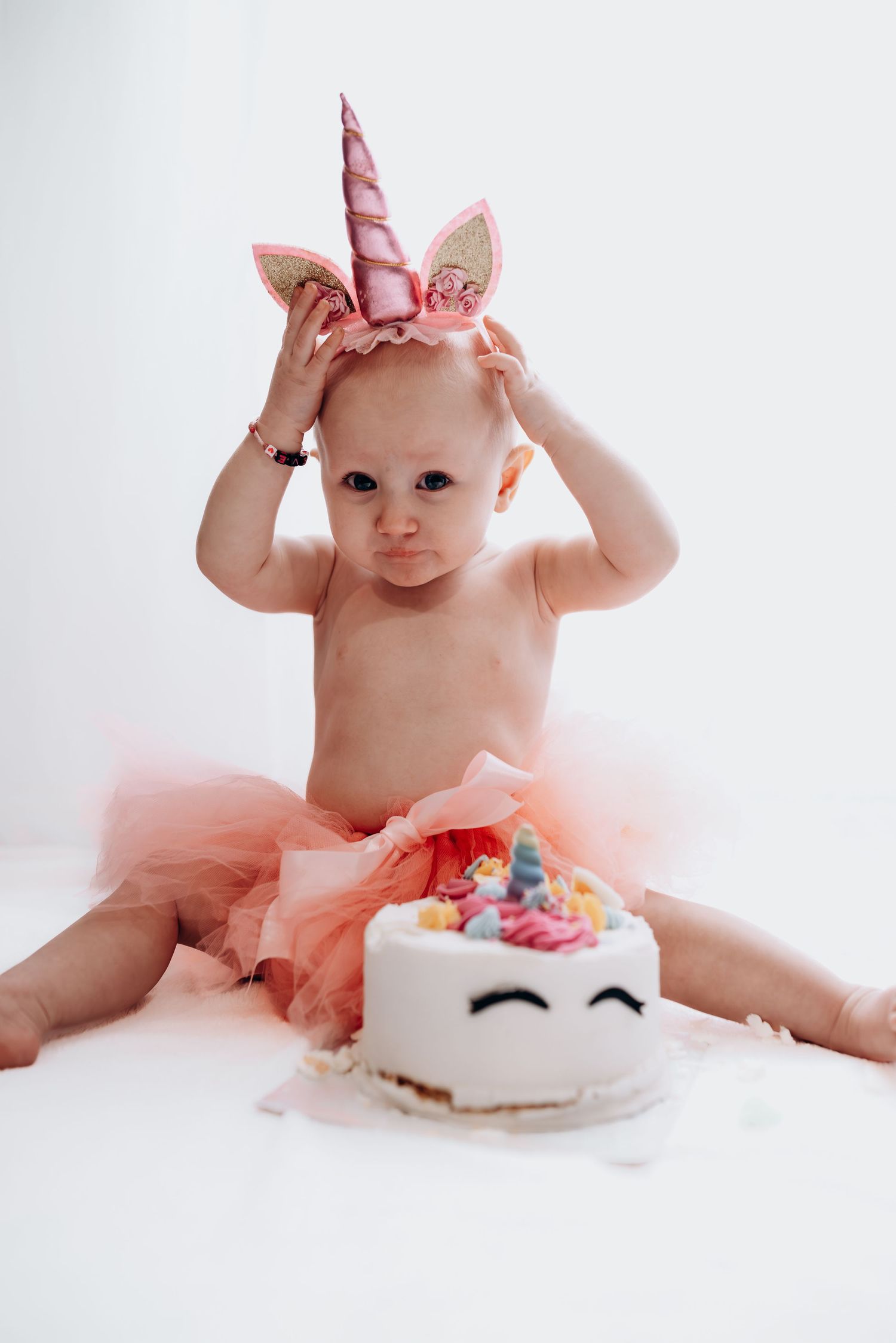 Baby in pink tutu exploring birthday cake during professional photoshoot against white background.