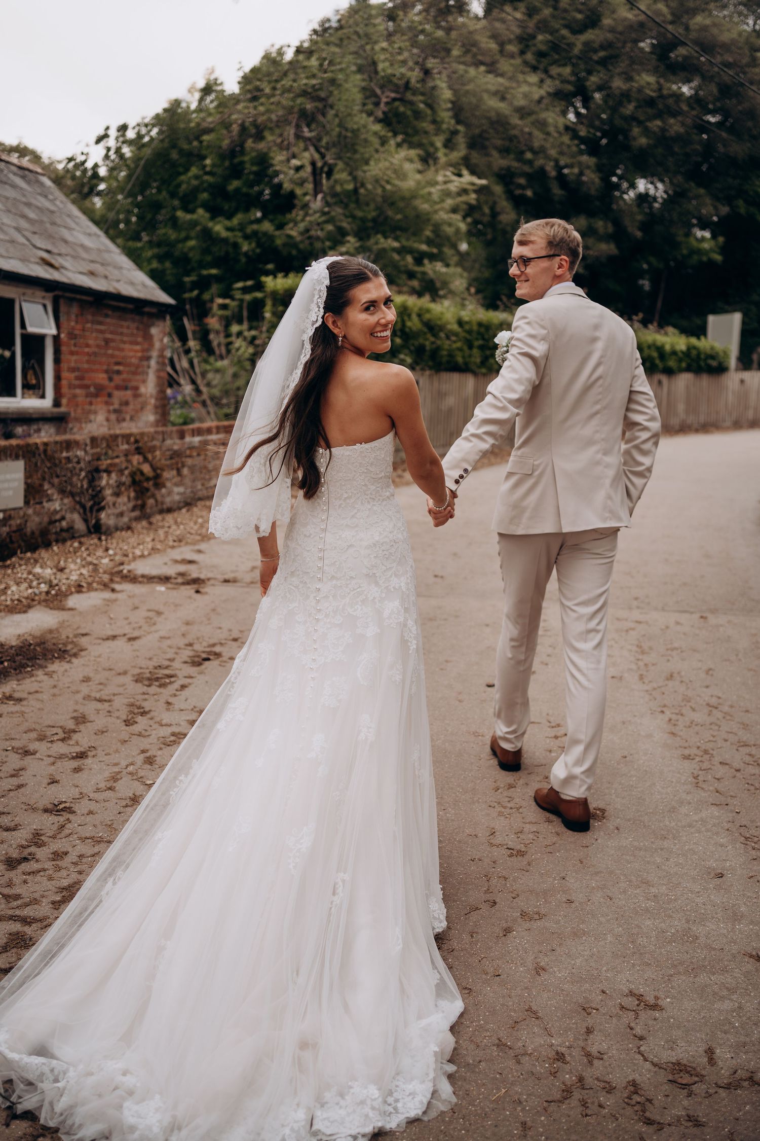 Bride in white lace gown and veil holds groom's hand walking on sandy path, smiling back at camera.