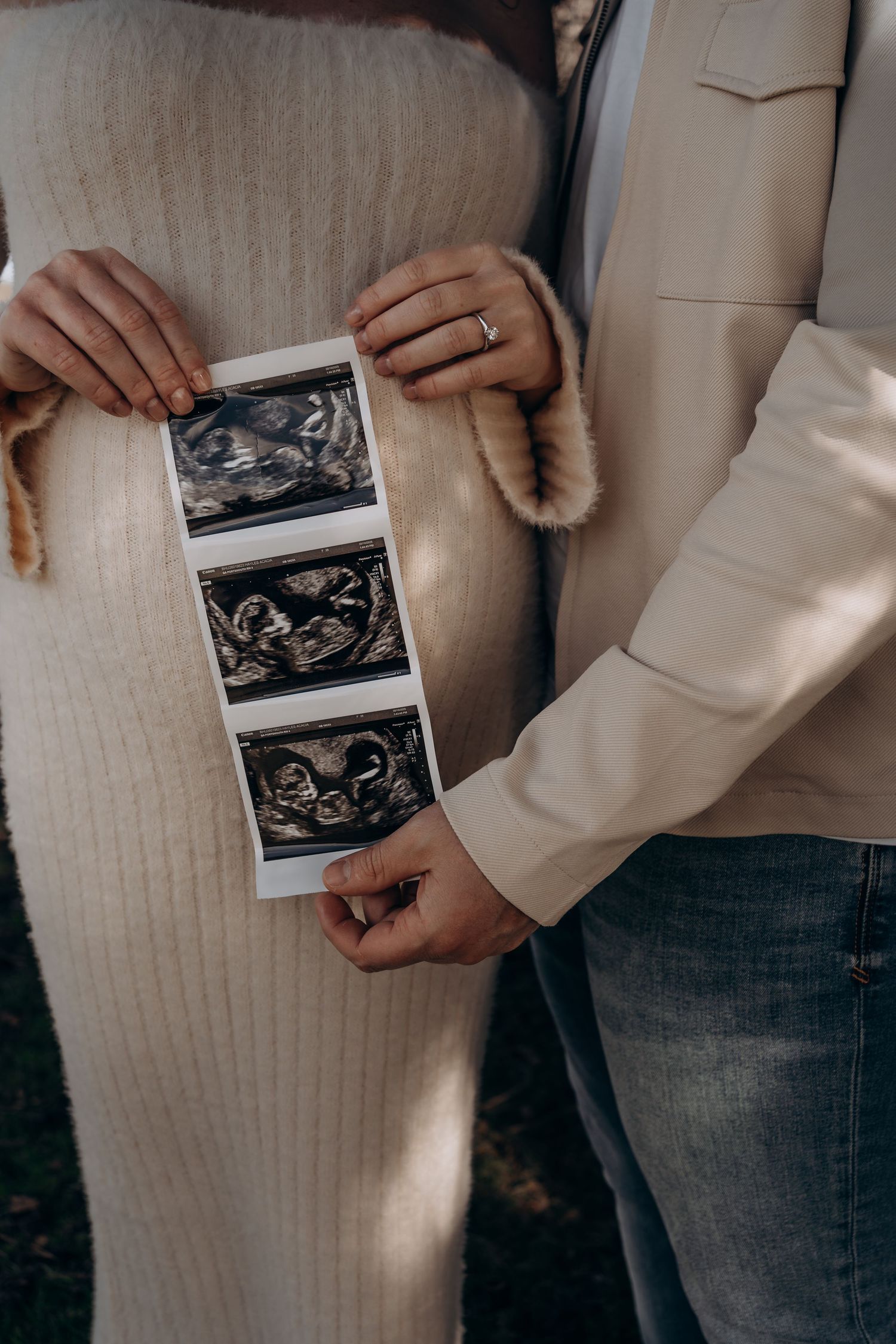 Pregnant couple holding ultrasound sonogram photos in front of baby bump, announcing pregnancy outdoors.
