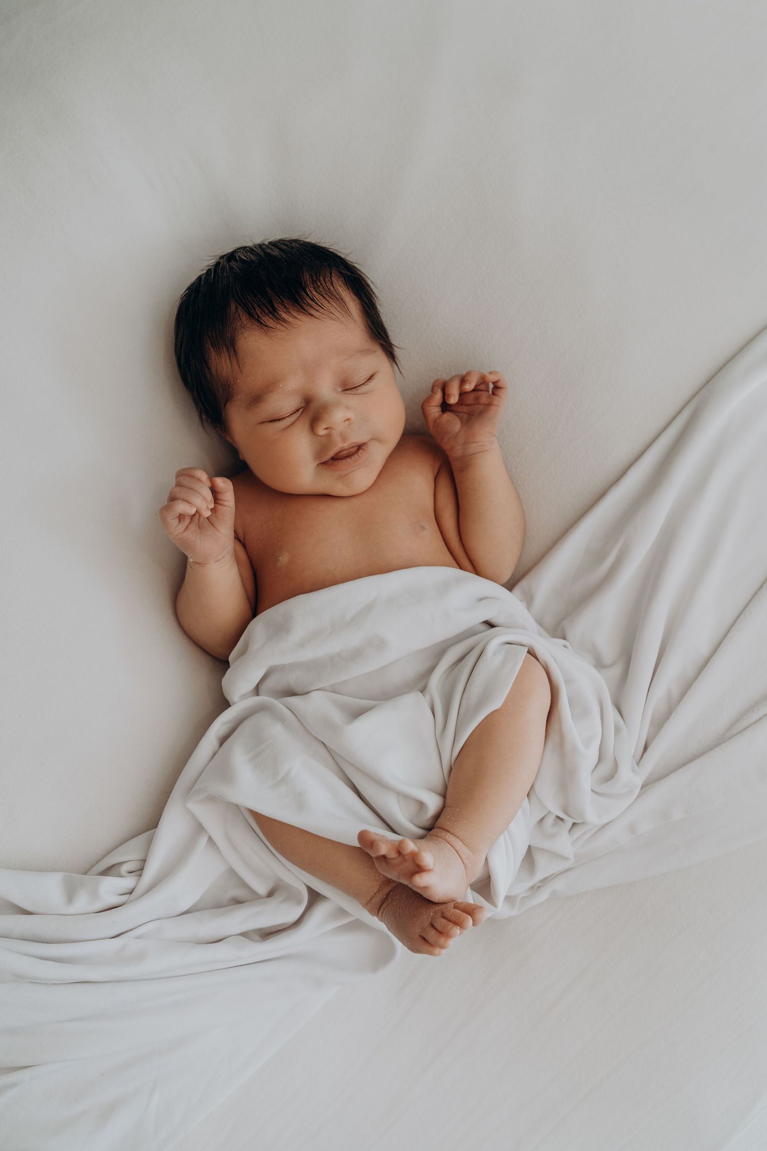 Peaceful newborn baby sleeping on white blanket with dark hair and tiny fists raised, captured in soft natural light.
