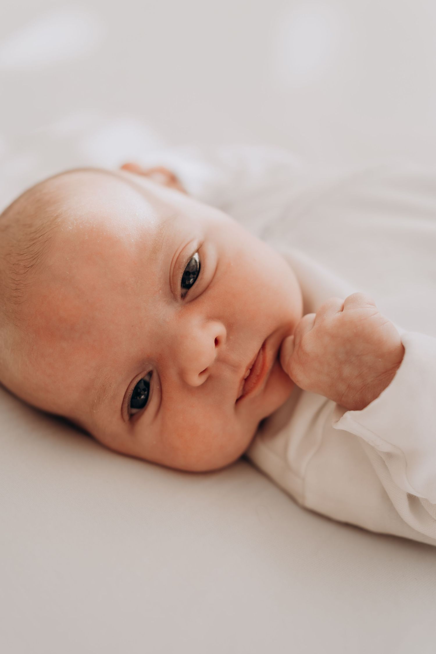 Close-up of infant lying on white bedding looking at camera.