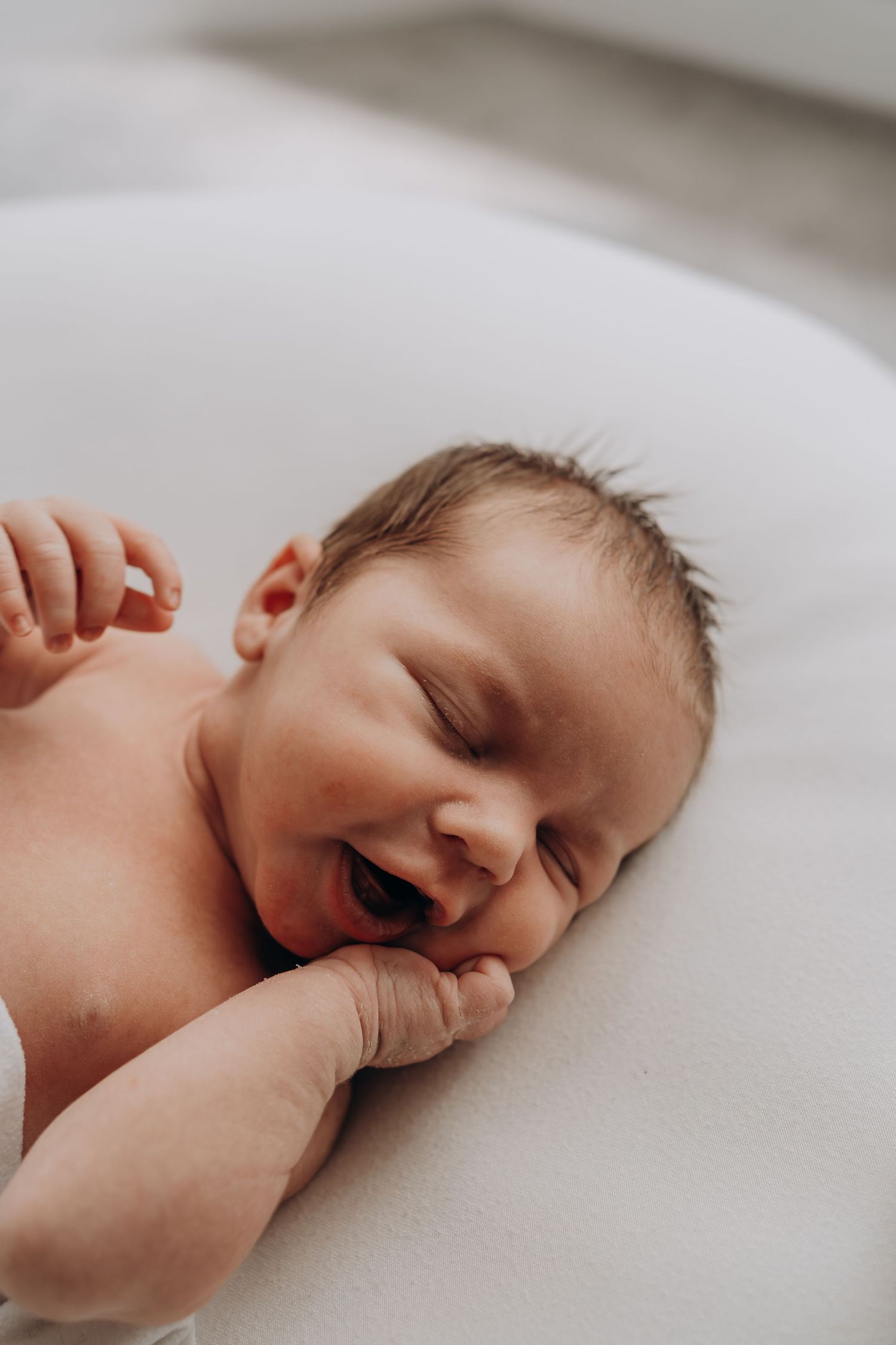 Newborn baby lying on white surface, eyes closed, mouth open, with tiny hands raised near face.