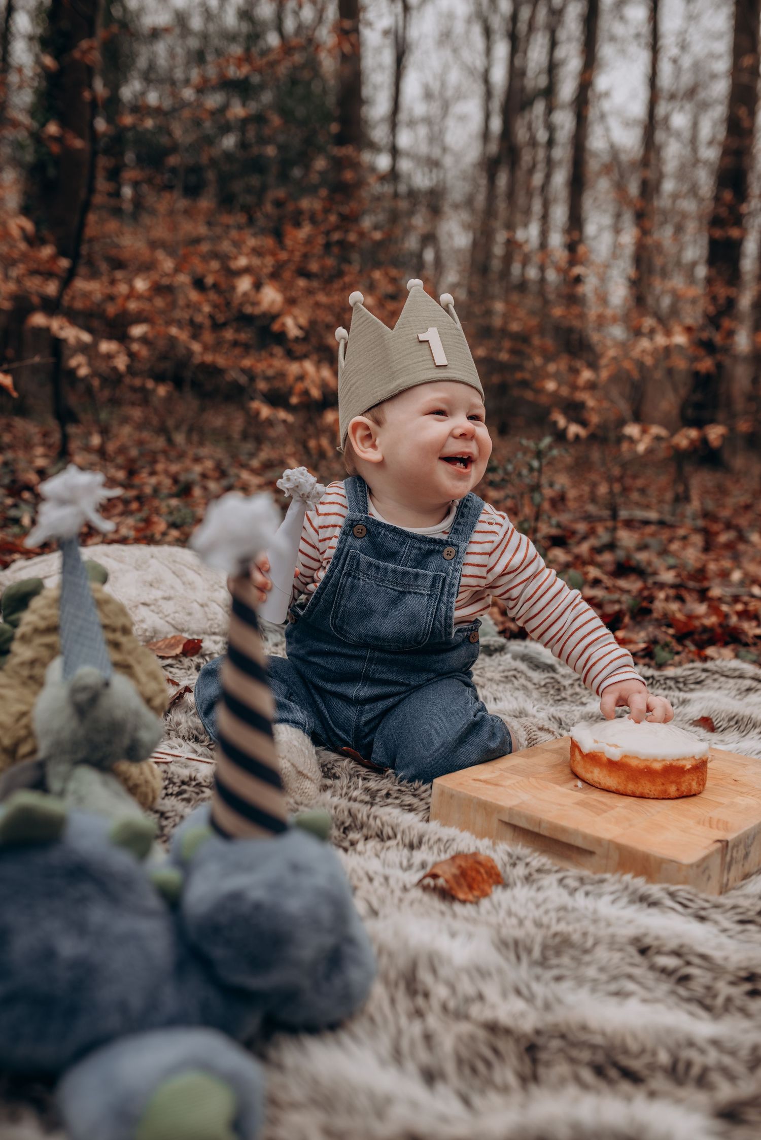 Baby in overalls and crown laughs during outdoor autumn cake smash photography session.