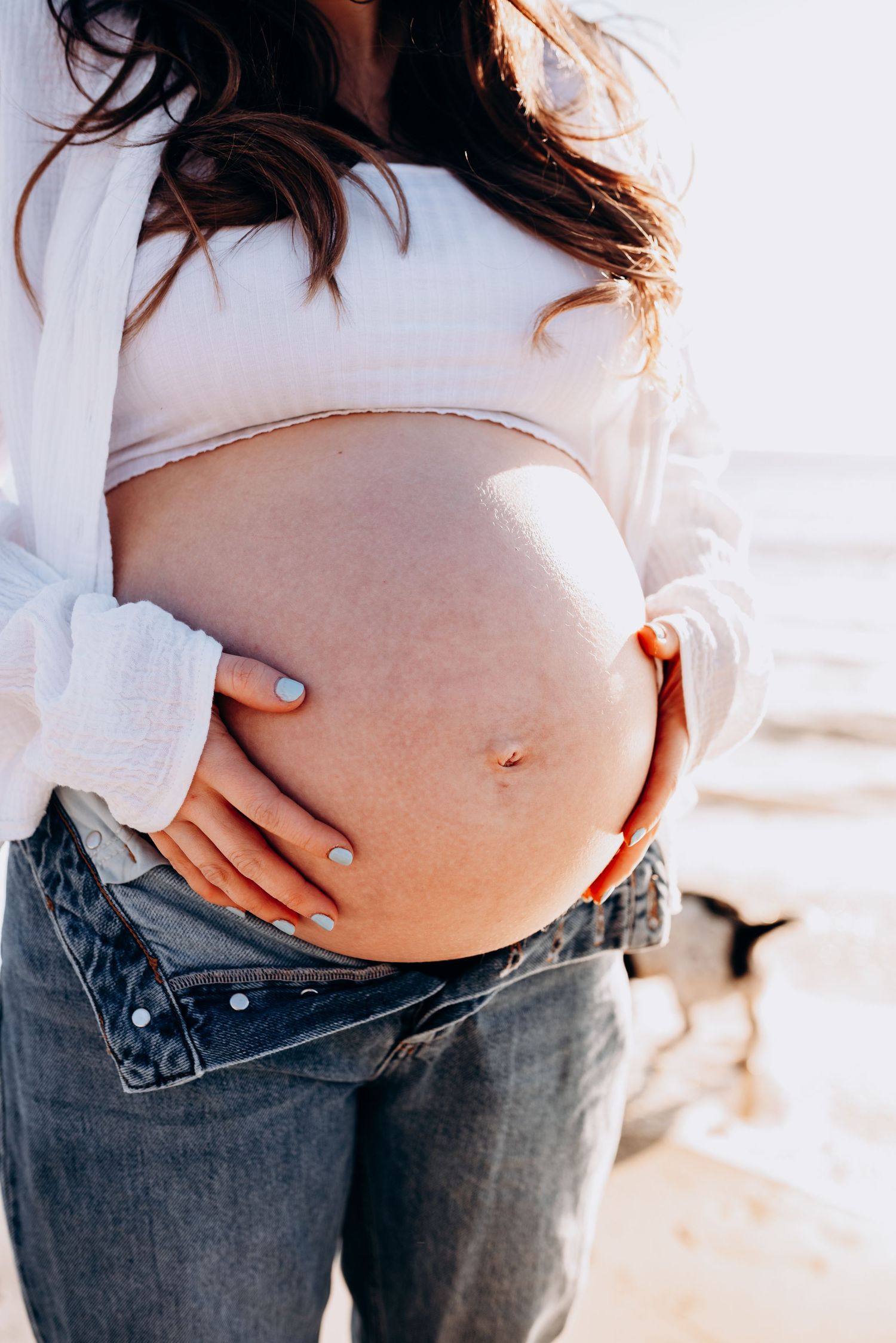 Close-up of pregnant belly with hands gently cradling bump against beach backdrop.