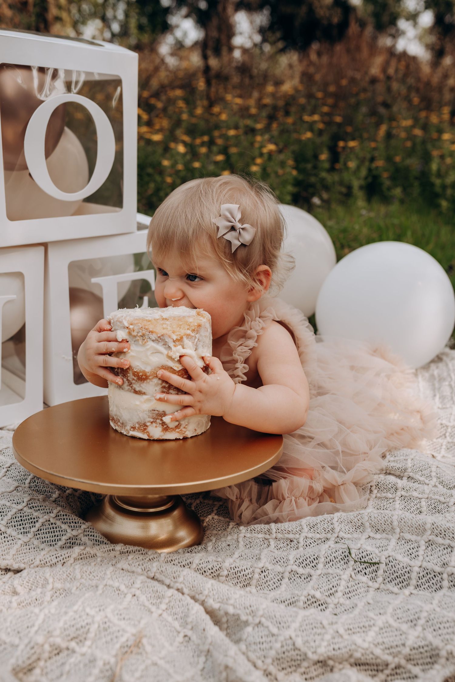 A baby in pink tulle dress eagerly bites into a birthday cake during outdoor photoshoot.