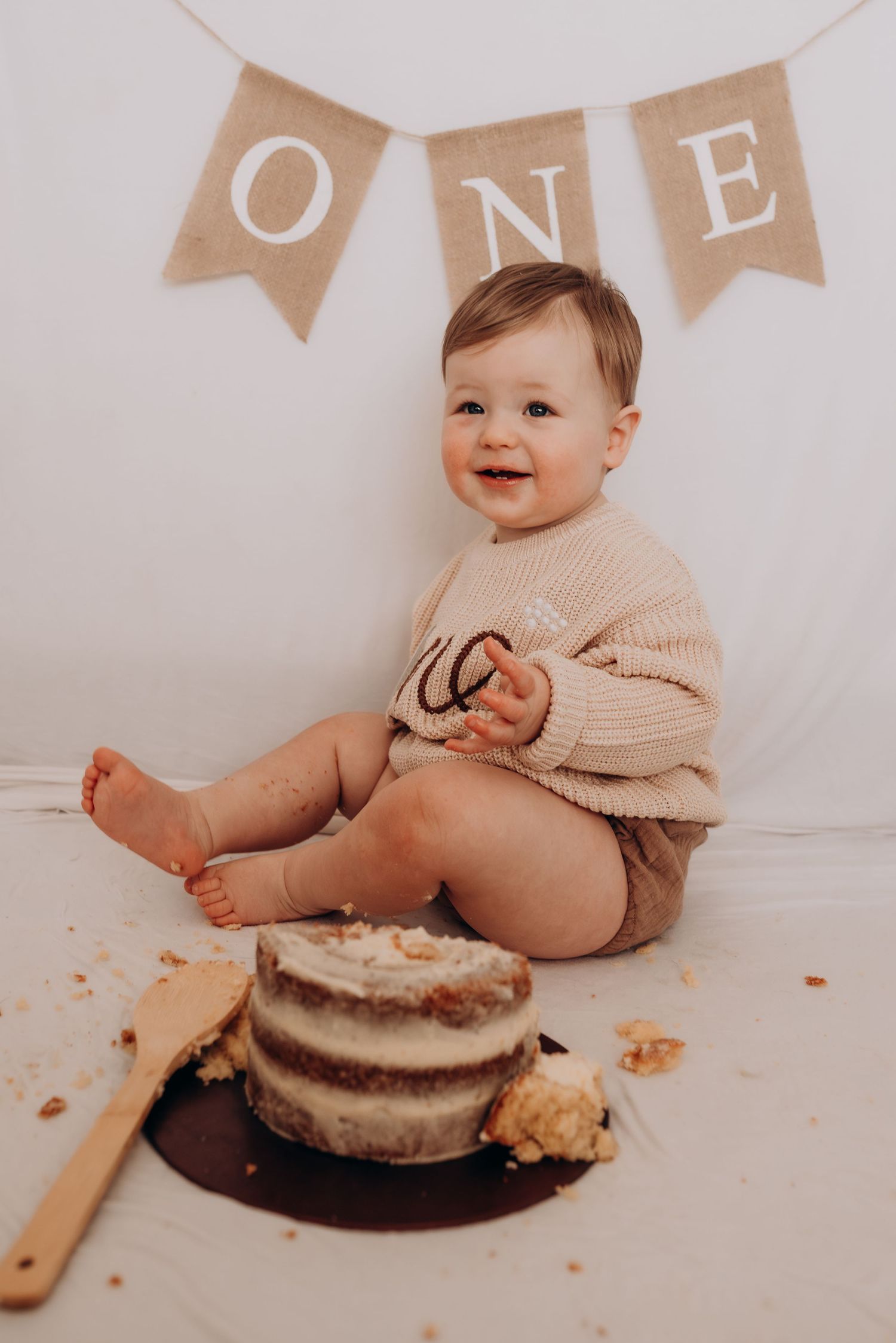 Baby wearing beige outfit sits on white floor with cake during first birthday photoshoot.