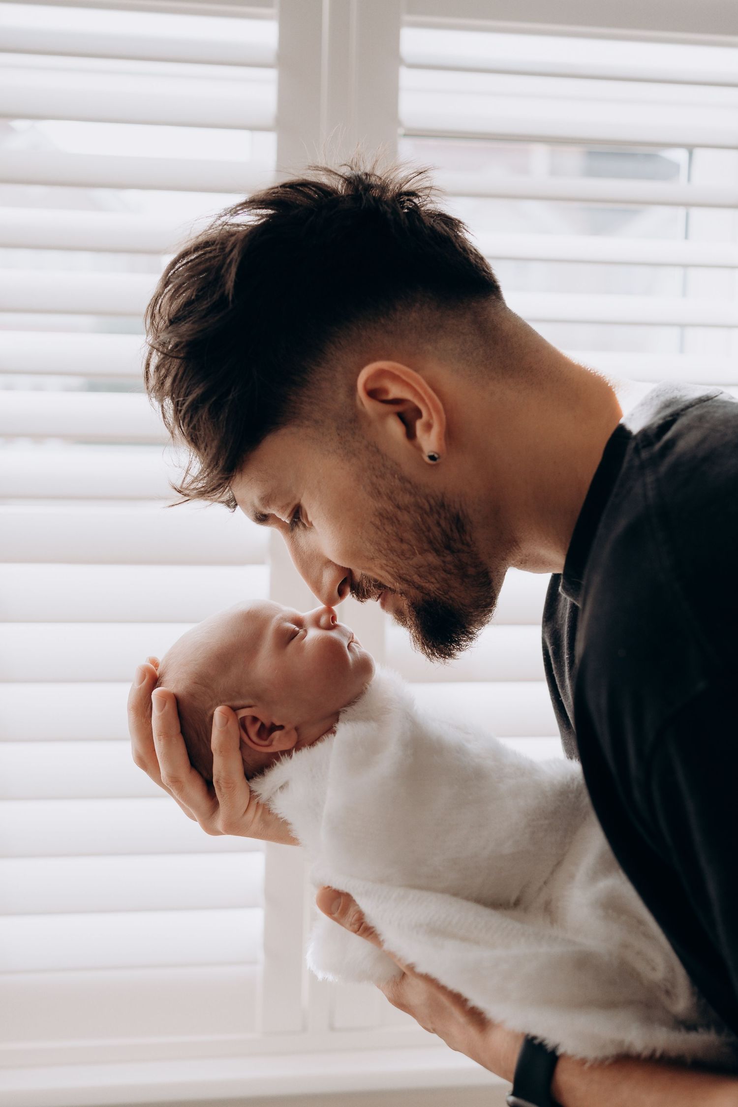 Young bearded father gently kissing his newborn baby wrapped in white blanket near window with blinds.