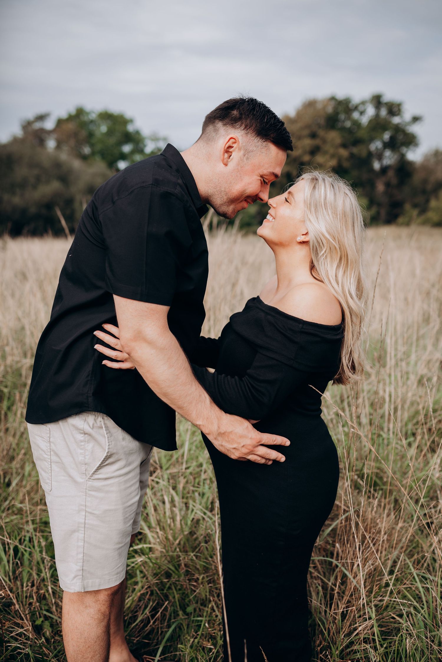 Intimate maternity portrait of couple in black attire sharing tender moment in grassy field.