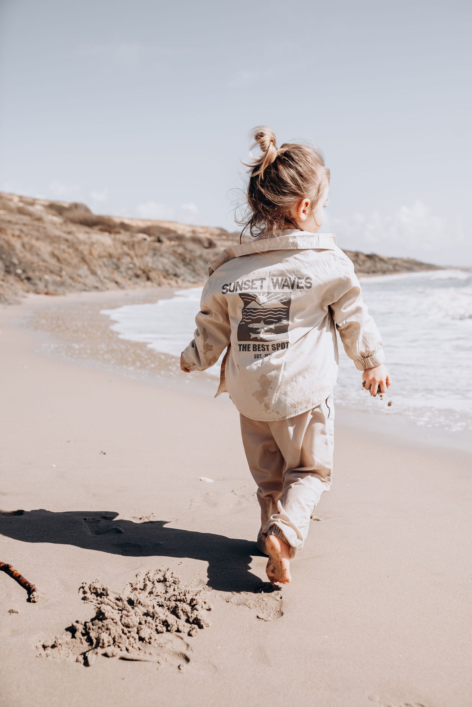 Child running and playing in sand on sunny beach wearing beige jacket.