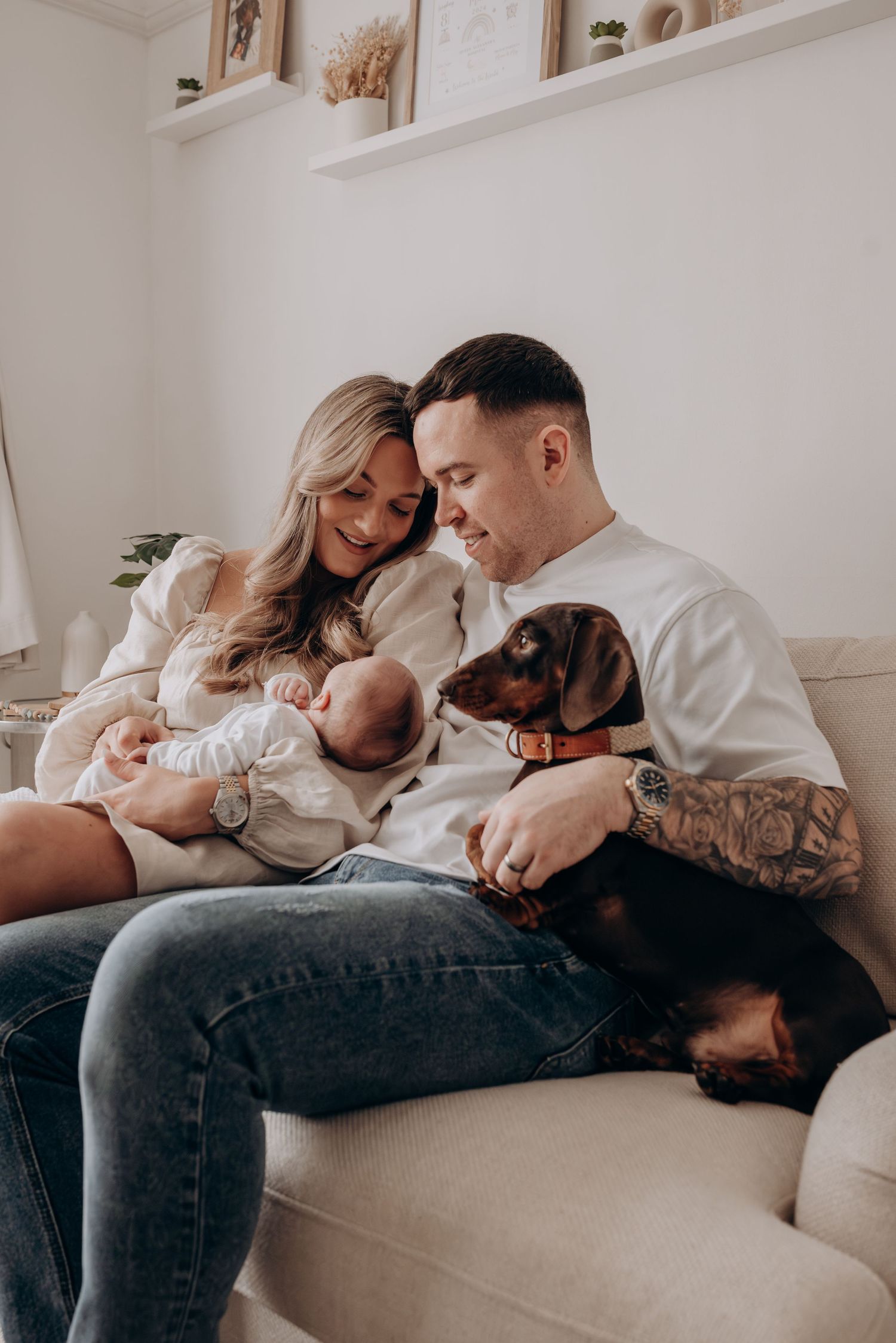 Family lounging on beige couch with newborn baby in casual intimate setting.