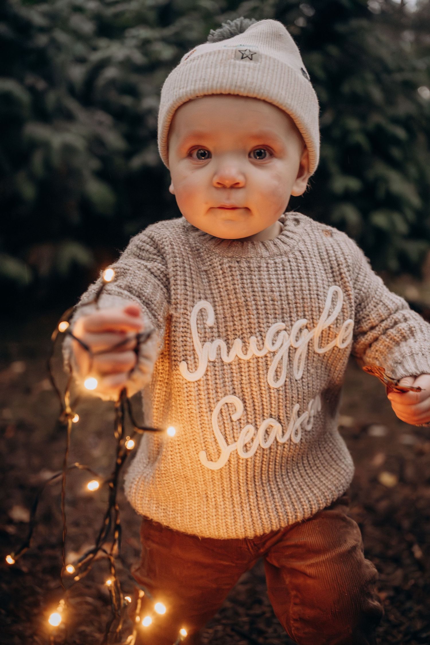 A small child in a gray knit sweater and beanie holds glowing string lights while standing outdoors at dusk.