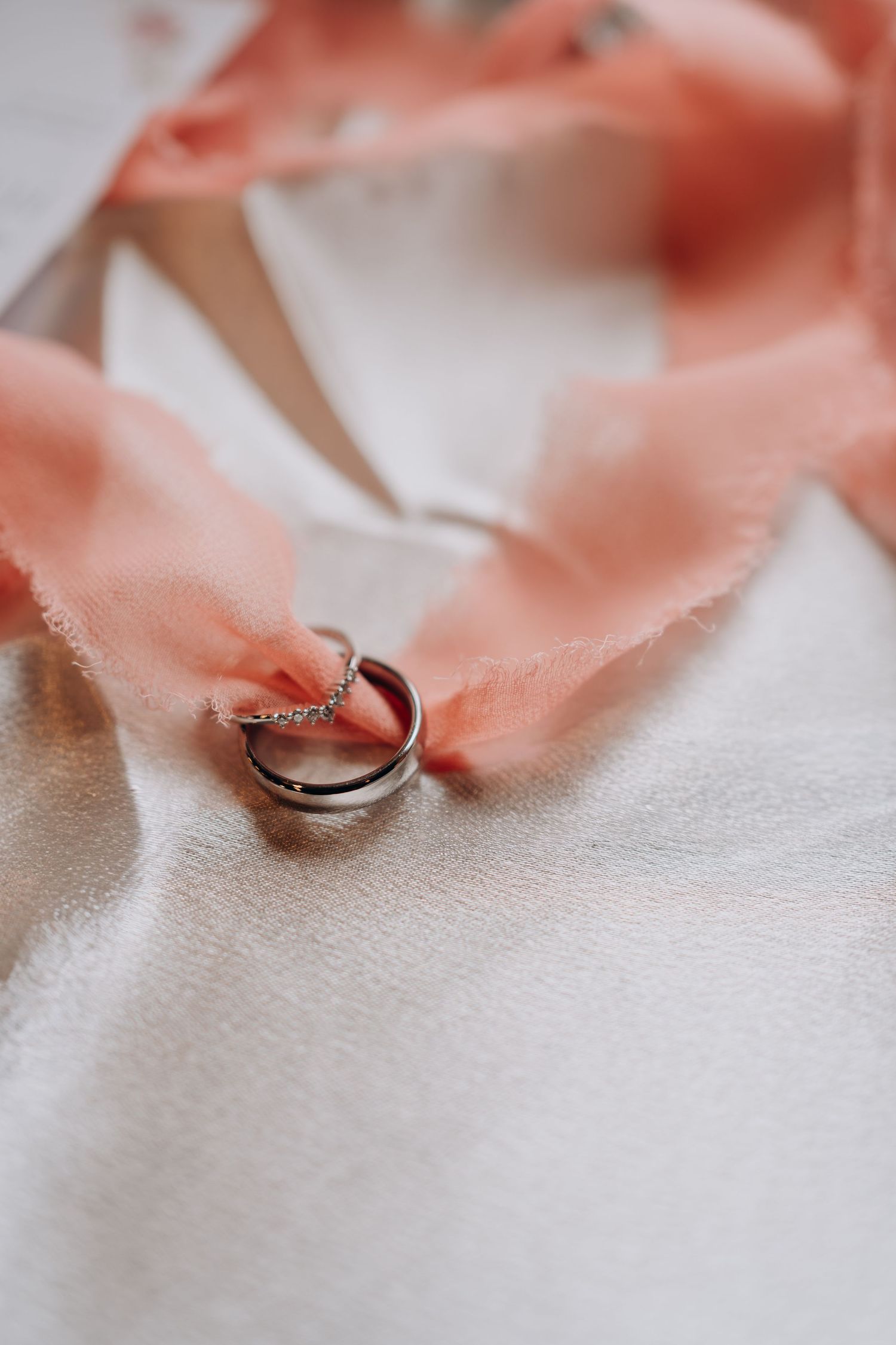 Wedding rings rest on a soft white surface with delicate coral-colored silk ribbon creating an elegant composition.