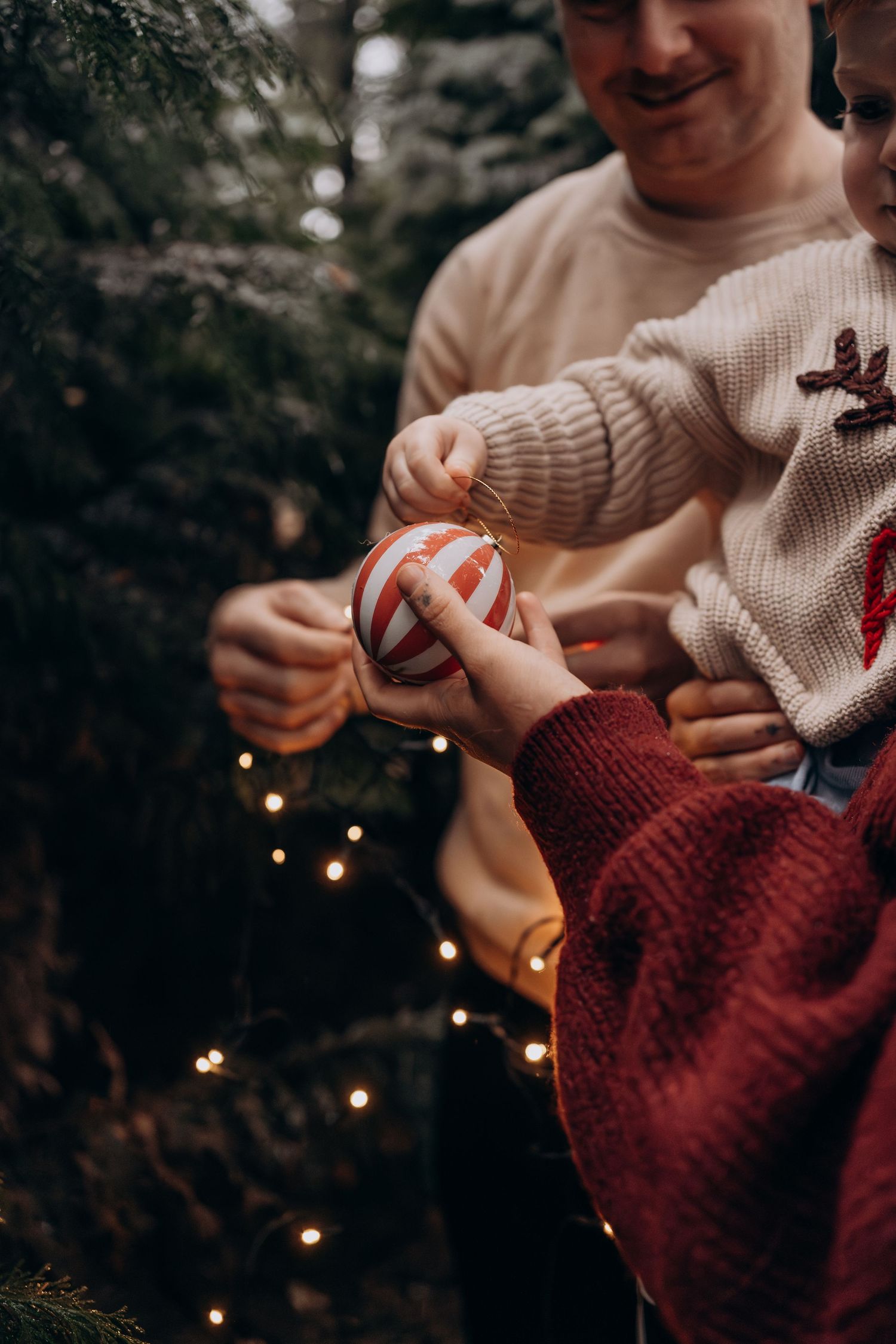 Hands holding a striped Christmas ornament near a decorated tree with twinkling lights in a cozy holiday setting.