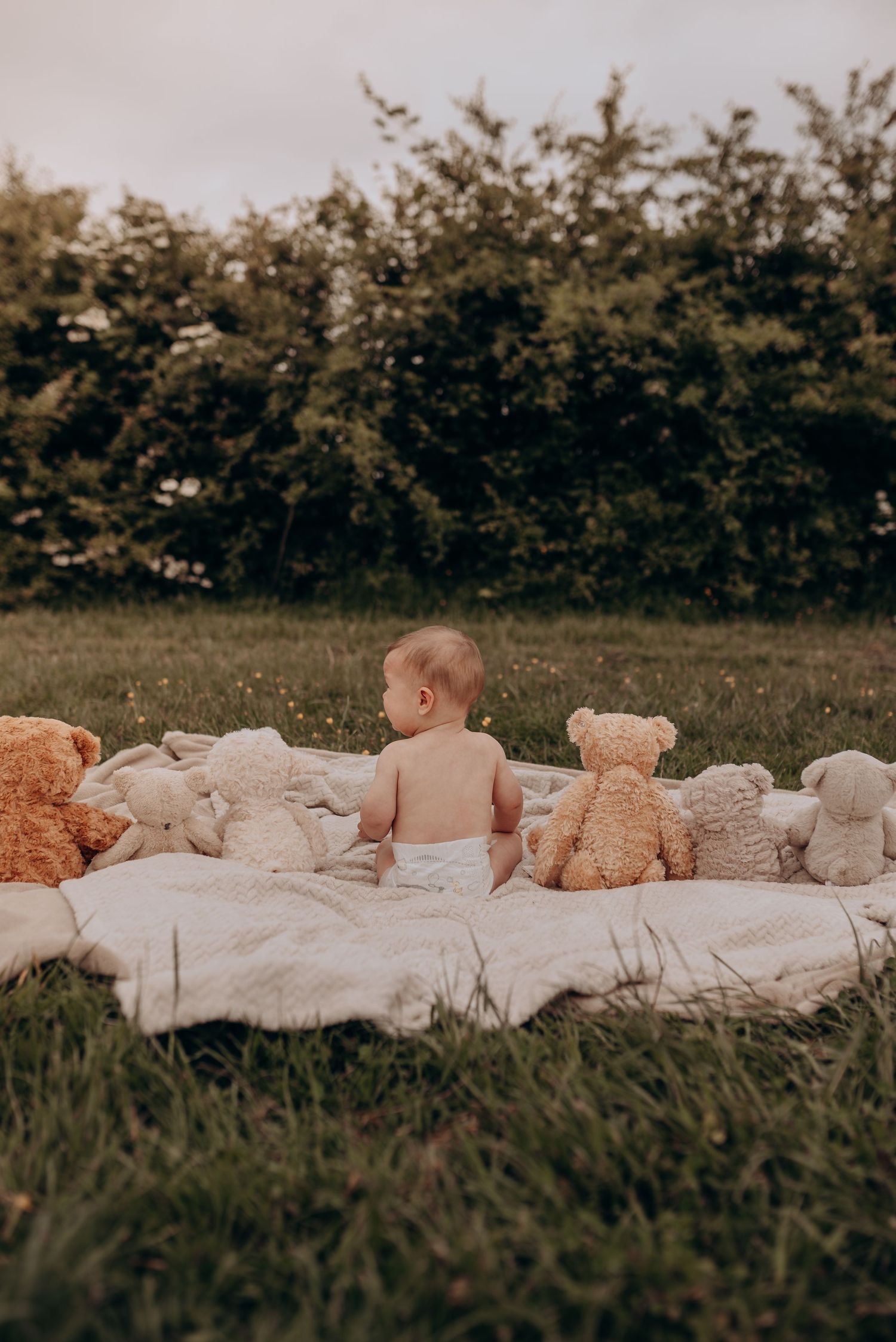 A baby lies on a white blanket surrounded by plush teddy bears in an outdoor grassy setting.