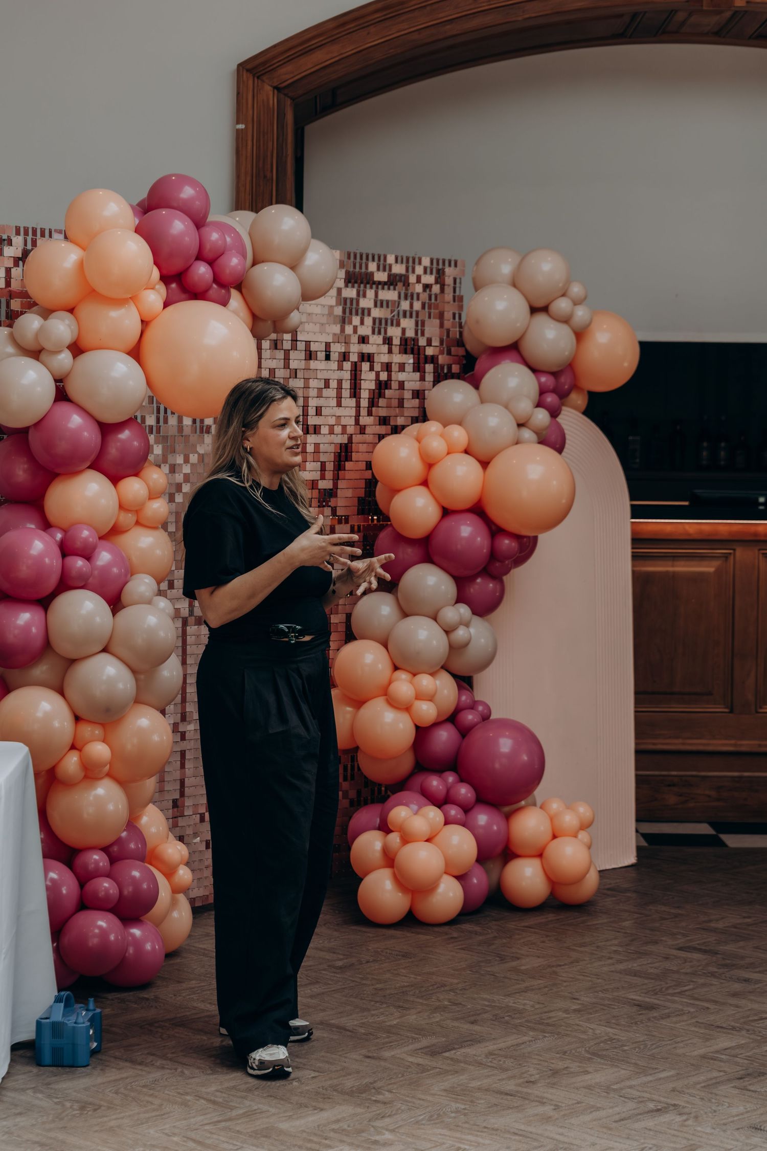 Woman in black outfit stands before colorful pink, orange, and beige balloon arch garland at festive event.